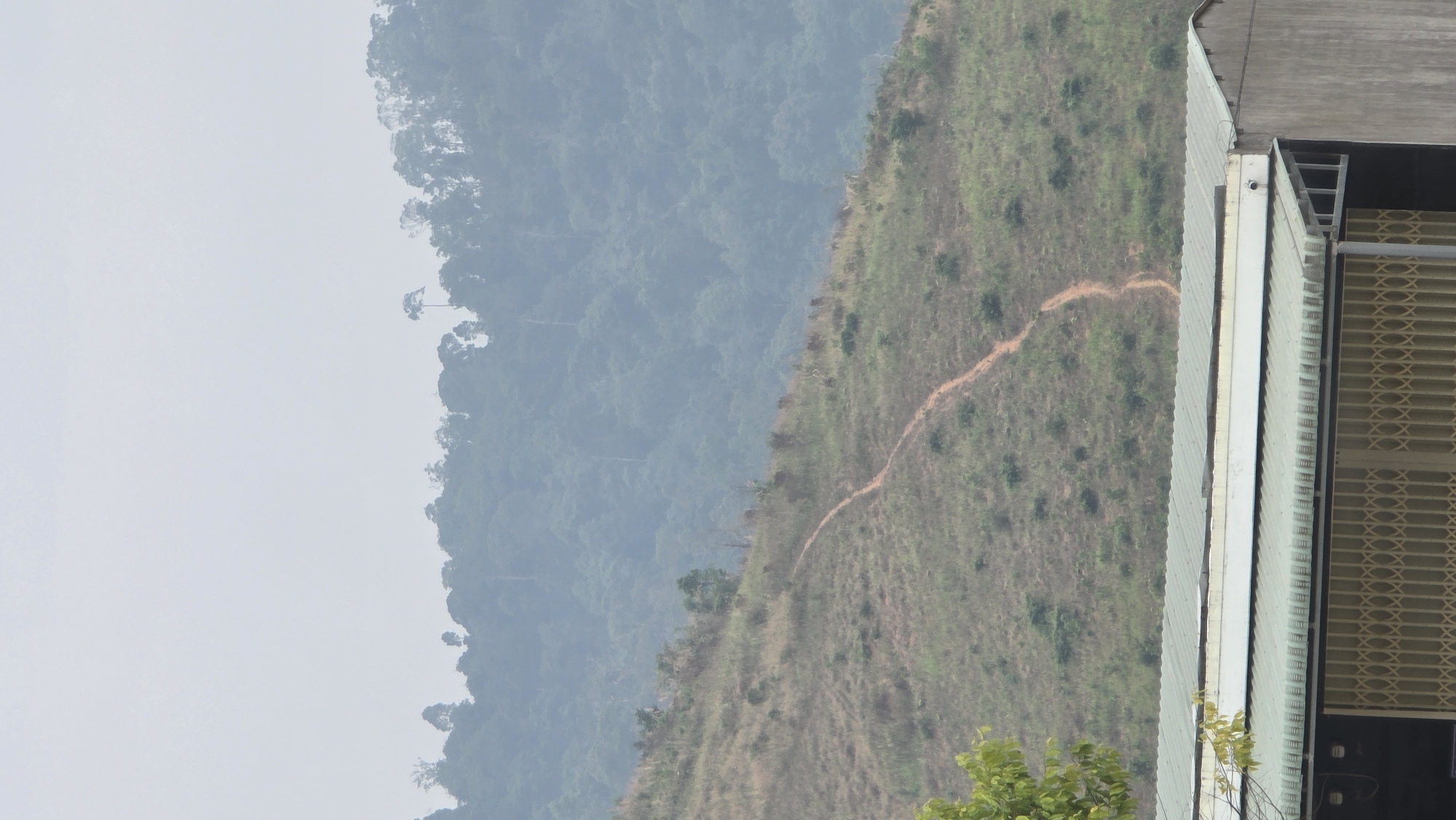A landscape shows a hillside bordering a tall building. A tan dirt trail winds diagonally up the right side of the hillside. To the left of the hillside, a hazy mountain range is visible, its texture suggestive of dense forest. To the right of the hill is part of a building. The building's walls are light grey, with some darker areas and grill-like sections. At the bottom right of the image are green leafy branches. The sky in the background is light grey. The picture was taken in Bo Y, Vietnam.