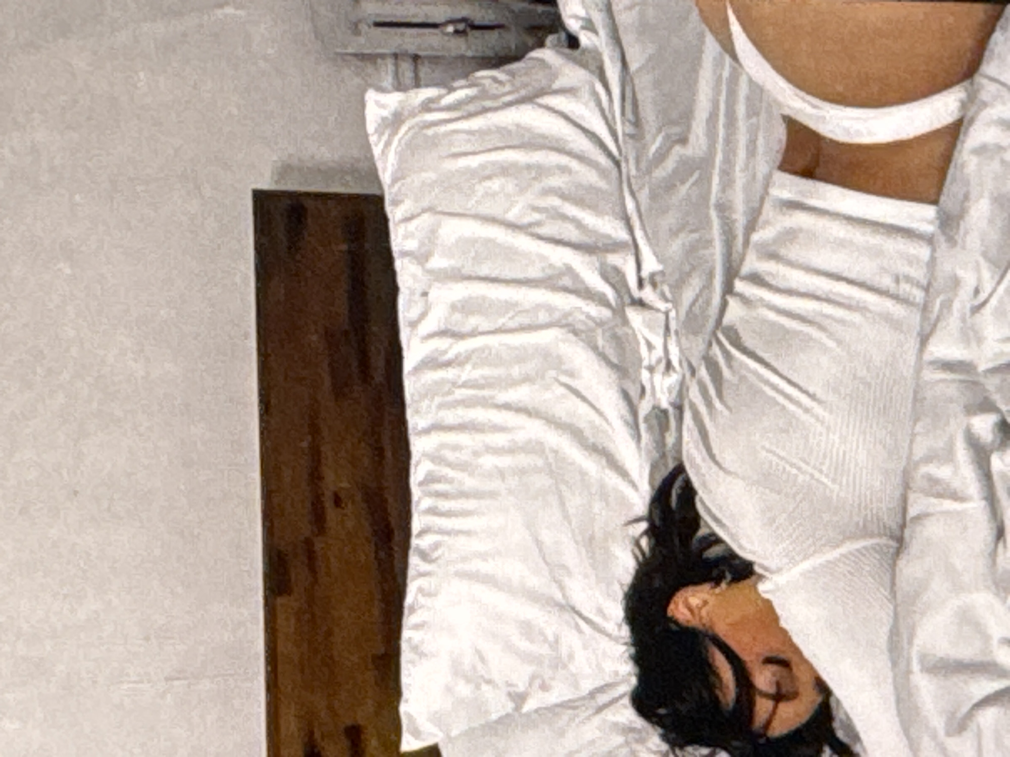 A person with dark hair is lying face down on a bed. The person is wearing a white long-sleeved top and white underwear. The bed has white sheets and a large white pillow. A wooden headboard is visible behind the pillow. The environment appears to be a bedroom. The lighting is soft and diffused. No specific location in Kuta Selatan, Indonesia can be determined from the image.