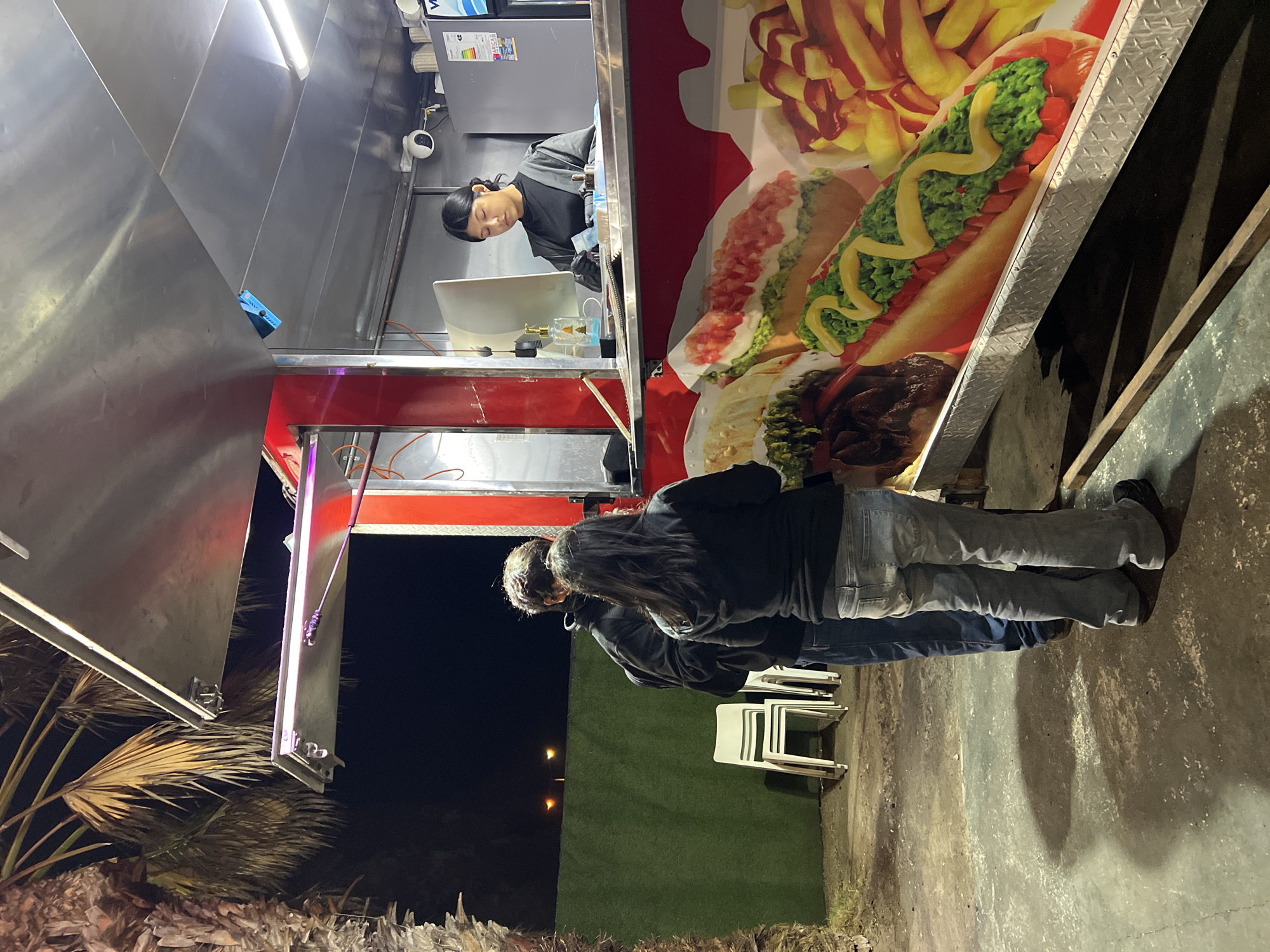 The image shows a food truck in Arica, Chile. A woman is working inside the food truck, serving two customers who are standing outside. The food truck has images of hot dogs and fries on its side. Stacks of white chairs are placed beside a green wall, on a concrete surface. A palm tree is visible on the left, under the night sky.