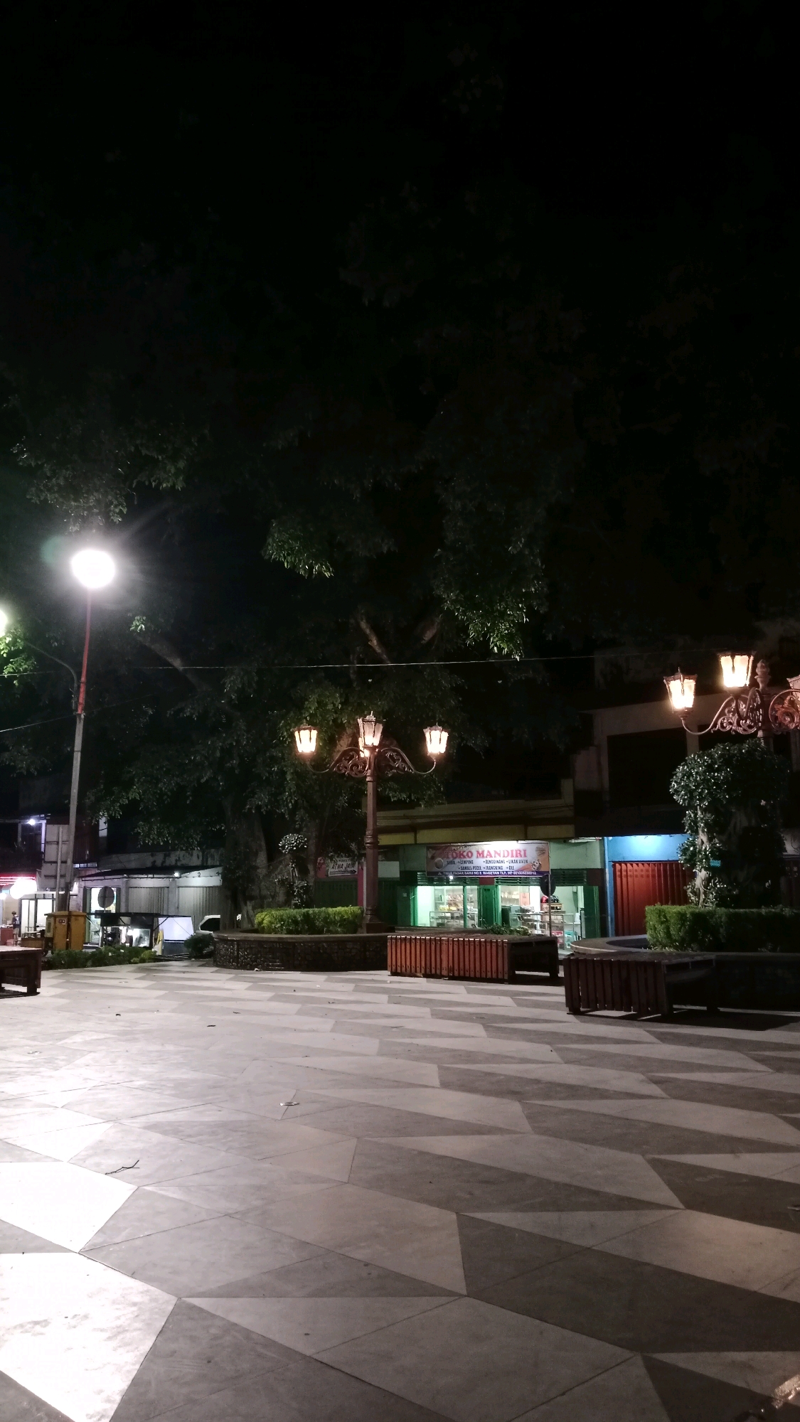 The image shows a night-time view of an outdoor public space in Magetan, Indonesia. The foreground is a paved plaza with a geometric pattern of light and dark gray tiles. In the mid-ground, there are several elements including: a decorative lamppost with three glowing yellow lights, a circular planter with green shrubs, and a building with a sign that reads "TOKO MANDIRI". There are also wooden benches visible. In the background, there are trees with dark foliage and more buildings, one with a blue facade and red doors. A street lamp with a bright white light is visible on the left side of the image. The overall scene is illuminated by artificial lighting from the lamps and the building. There are no people visible in the image.