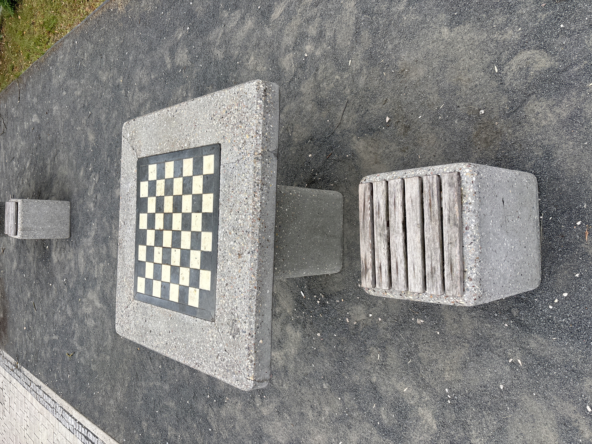 This is an eye-level, medium shot of an outdoor chess table and seating area. The chess table is made of concrete with a checkerboard pattern inlaid on its surface. In front of the table, there is a concrete bench with wooden slats. The ground is covered in dark gravel, with a paved area of grey and white tiles to the left. In the background, there is another concrete bench and some grass and foliage. The scene is set in Zvenigorod, Russia, likely in a park or public space. There are no people present.