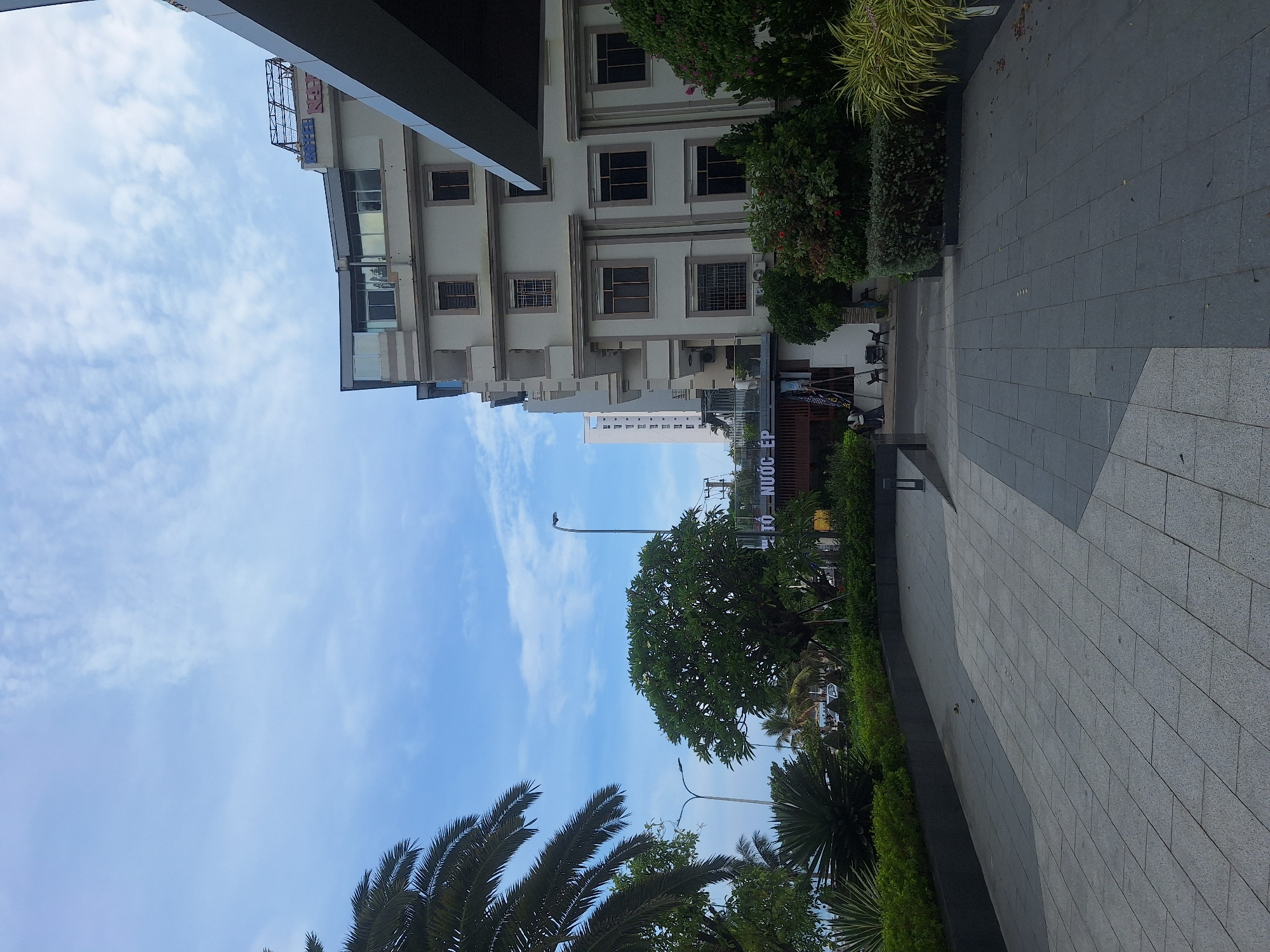 A street-level, eye-level, wide-angle view shows a modern, light-colored building with multiple floors and balconies, situated in Nha Trang, Vietnam. The building has numerous windows with dark frames and some with grates. The facade is accented with darker horizontal bands. A sign reading "TO NUOC EP" is visible on a lower section of the building, above a covered walkway. To the left of the building, a palm tree frond is visible in the foreground, with lush green landscaping and other trees, including a large tree with white flowers, and a decorative palm in the middle ground. A paved walkway, made of light grey square tiles, extends from the foreground into the scene. The sky is blue with scattered white clouds. A street light is visible between the palm tree and the building. In the background, other buildings and distant trees are visible. Some people are seen on ladders near the building's entrance, possibly engaged in maintenance or construction.