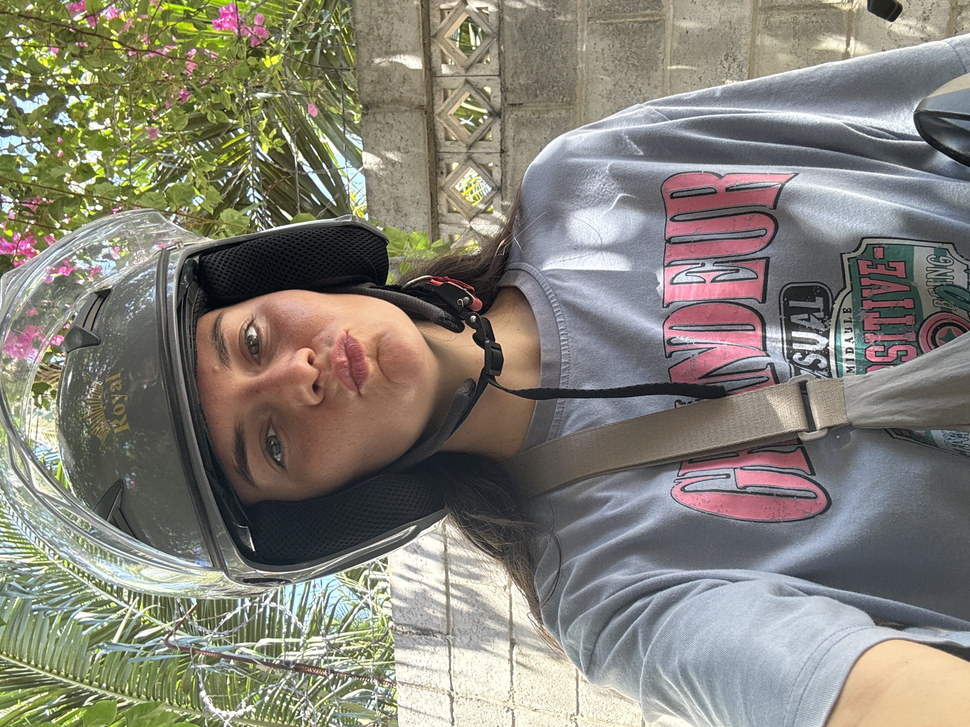 A person wearing a helmet and a grey t-shirt is looking at the camera. The helmet is a black and clear full-face helmet with "Royal" branding. The person has brown hair and is making a pouting face. A tan strap crosses the person's torso. The background includes a grey brick wall, green foliage, and pink flowers. The location is likely Mui Ne, Vietnam.
