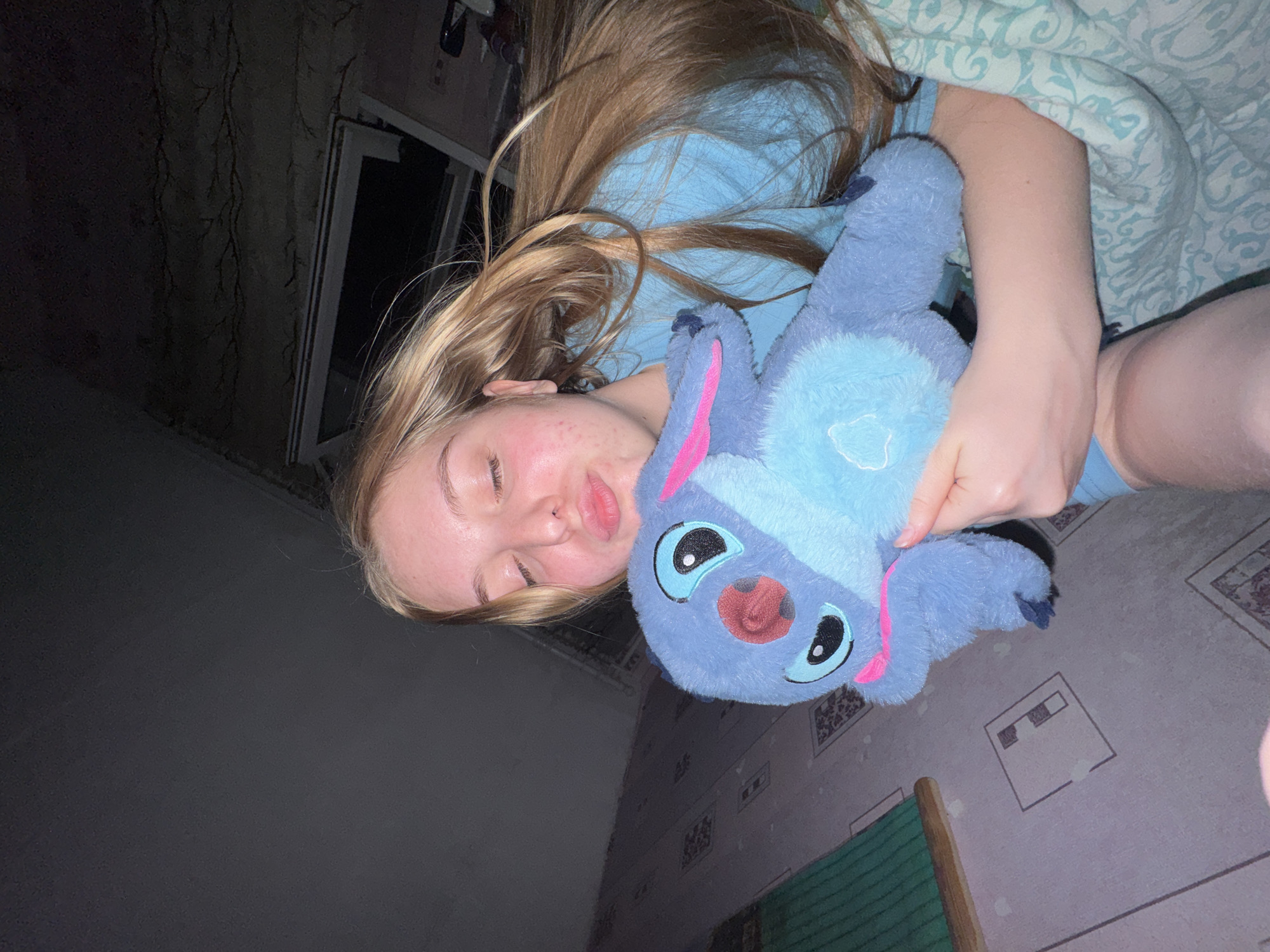 A fair-skinned person with closed eyes and blonde hair lies on a bed, cradling a blue plush toy resembling Stitch. The person is wearing a light blue shirt. The bed is covered with a patterned blanket. The background shows a dark room with a window and patterned wallpaper. The exact location within Perm, Russia, is not discernible from the image.