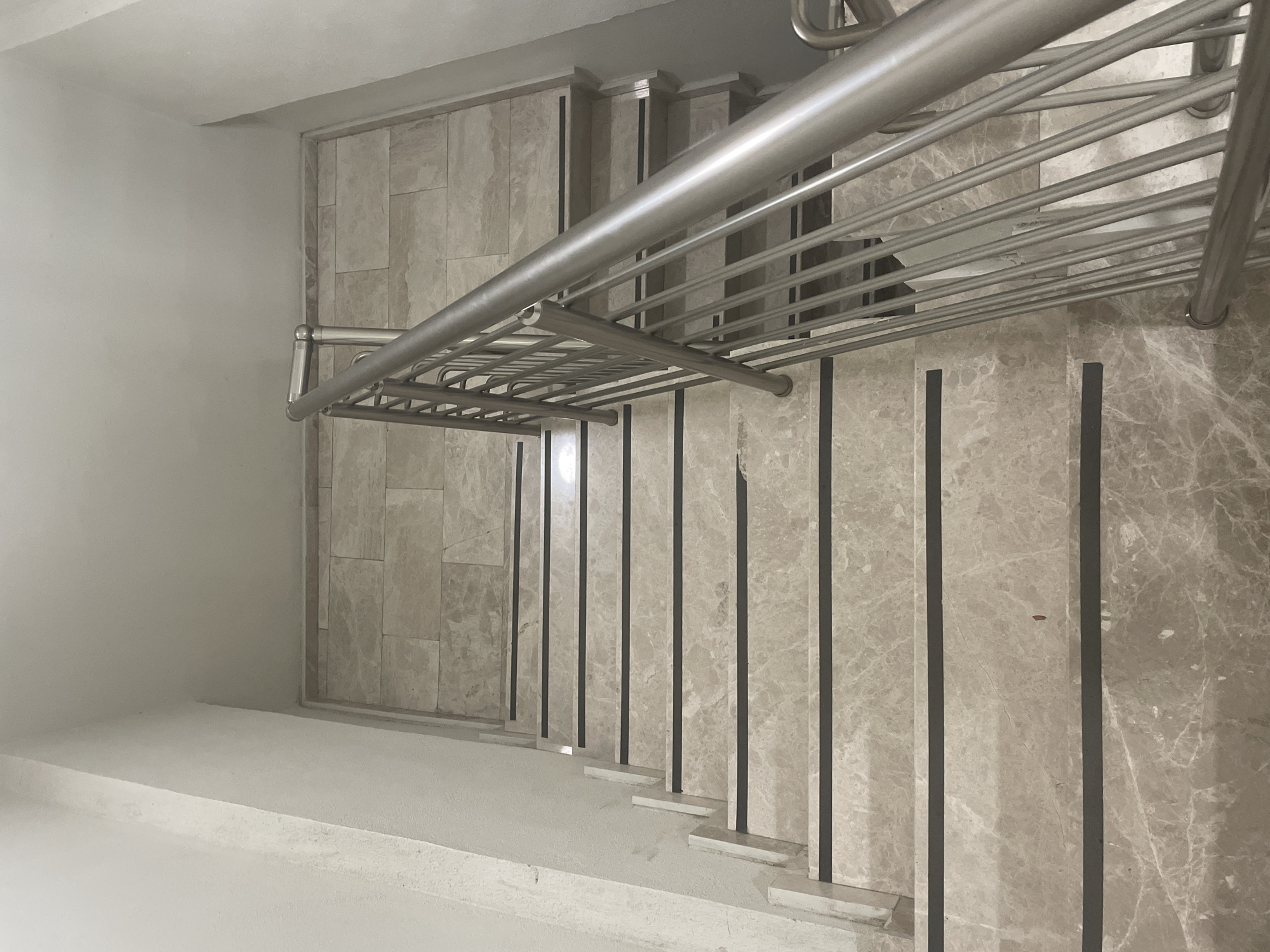 This is a high-angle, eye-level shot of a modern, marble-tiled staircase with a sleek metal railing. The staircase appears to be located indoors, possibly in a residential building or a commercial space in Yunus Emre, Turkey. The marble tiles on the steps and walls are a light grey with subtle veining, creating a sophisticated and clean aesthetic. Each step has a dark grey or black accent strip, providing contrast and likely serving as a non-slip feature. The metal railing is minimalist and consists of a thick handrail supported by numerous thinner horizontal bars, all in a brushed silver finish.
The lighting suggests it is daytime, with soft, diffused light illuminating the scene. There are no people or specific events visible; the image focuses entirely on the architectural details of the staircase. The surrounding walls are a plain, light color, likely painted. The overall impression is one of contemporary design and functionality. The image is well-composed, highlighting the geometric patterns of the tiles and the clean lines of the railing. There is no visible text in the image.
