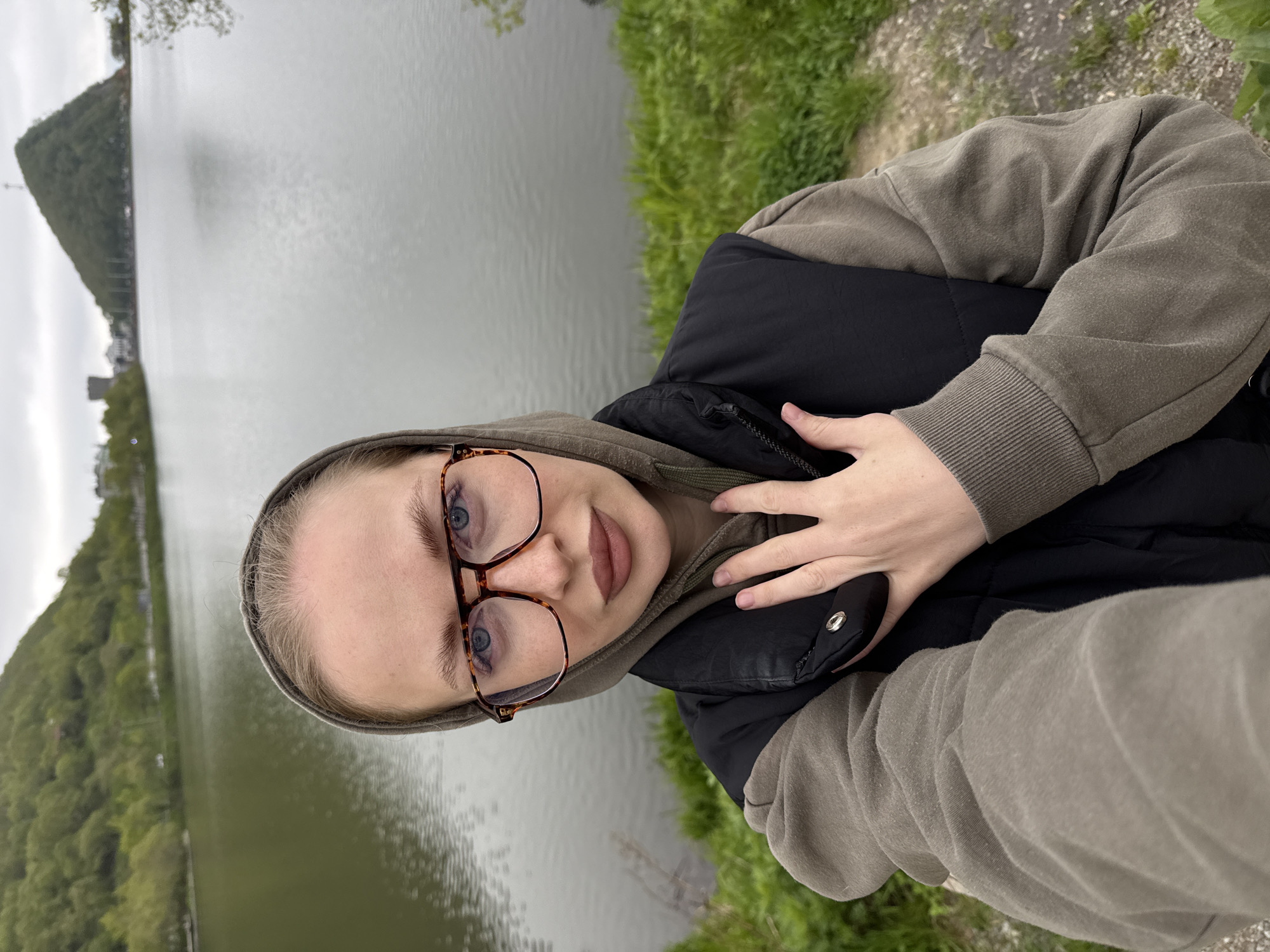 A woman with blonde hair and blue eyes, wearing glasses, a hoodie, and a vest, stands in front of a lake. The location is the city of Petropavlovsk-Kamchatsky, Russia. The background shows a hill with a cross on top, a tower, and trees. The sky is overcast.