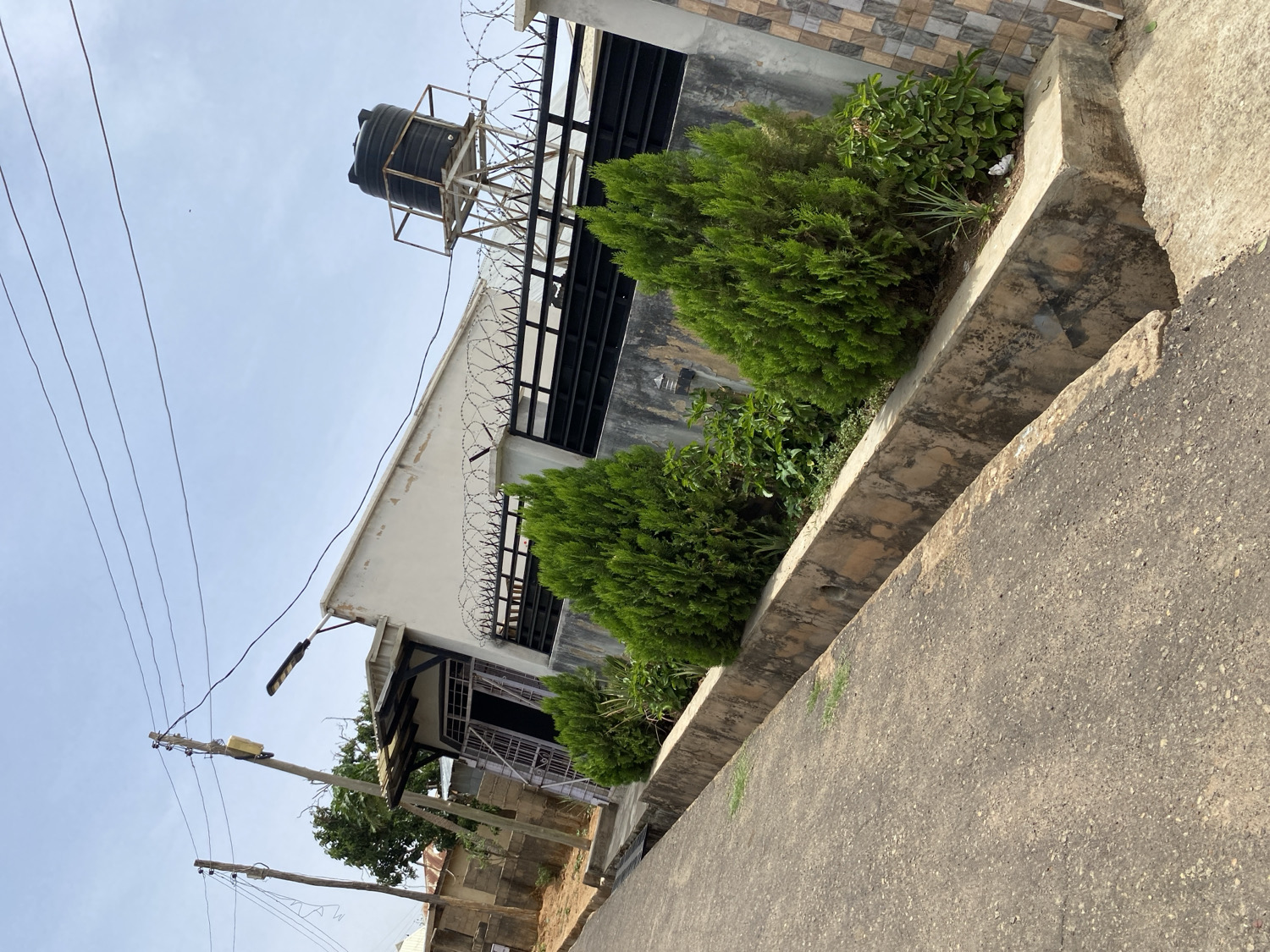 The image displays an outdoor scene, likely in a residential or commercial area in Jos, Nigeria, under clear daylight conditions.
On the right, a light-colored building with a flat roof is prominent. The top of its wall is secured with dark horizontal railings, topped with razor wire. Above the building's roof, a black plastic water tank is elevated on a metal stand, silhouetted against a bright, clear sky.
In the foreground and extending along the right side of the building, a raised concrete planter box contains various green plants, including several conical evergreen bushes. Adjacent to this planter is a light-colored concrete or asphalt road or pathway.
On the left side of the scene, two wooden utility poles stand vertically, supporting multiple overhead power lines that stretch across the sky. One of these poles is fitted with a street lamp and a small yellow rectangular box. The bright sky suggests it is late morning or early afternoon with sunny weather. No people are visible, and no specific activity is taking place. There is no discernible text in the image.
