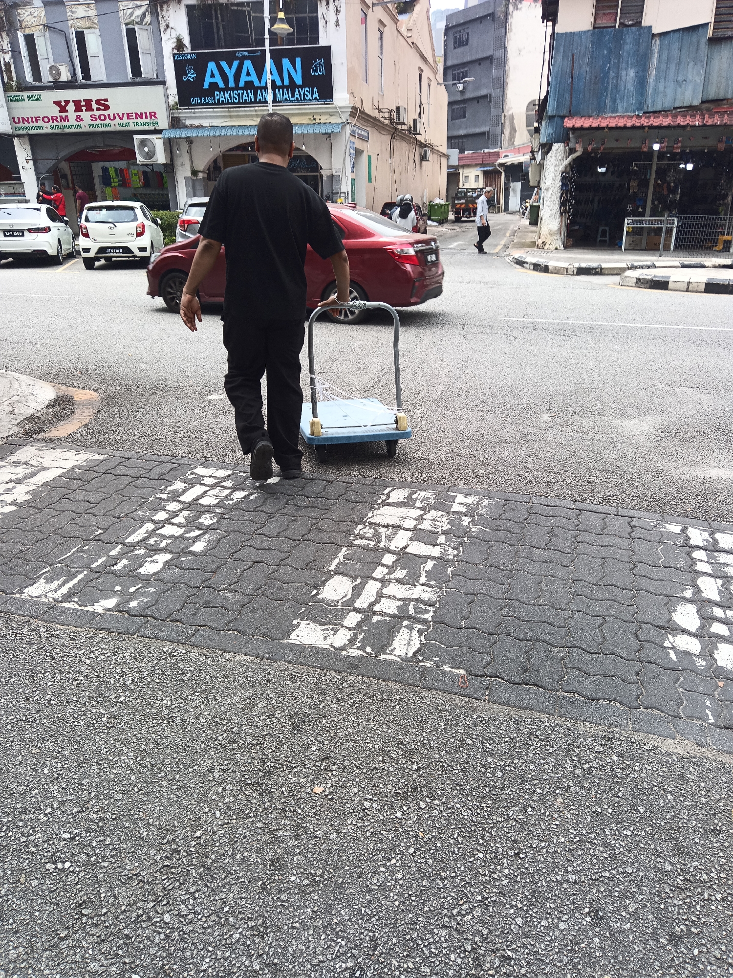 A full shot shows a man from behind pushing a blue cart across a pedestrian crossing. He is wearing a black t-shirt and pants and appears to be walking on a street in Kuala Lumpur, Malaysia, as indicated by the signage in the background, which includes "AYAAAN RESTORAN CITA RASA PAKISTAN AND MALAYSIA" and "YHS UNIFORM & SOUVENIR". The street is paved with asphalt and the pedestrian crossing is made of interlocking bricks with white stripes. Several cars are parked and driving on the street. The buildings in the background suggest a commercial area, with shops and businesses lining the street.