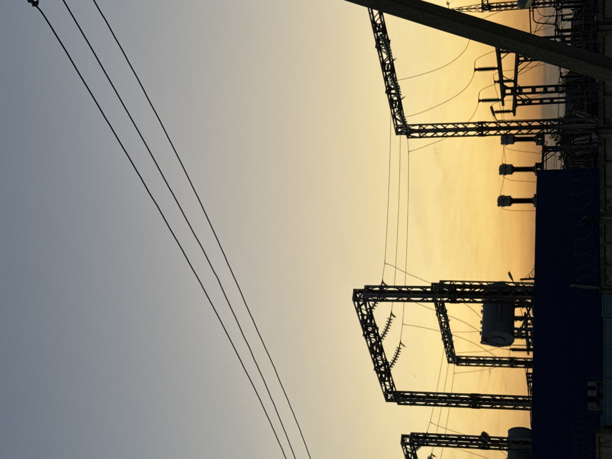 A wide shot captures a silhouette of electrical infrastructure at dusk. The sky transitions from a pale blue at the top to a warm orange and yellow hue near the horizon. Three thick power lines stretch diagonally across the upper portion of the frame. Below them, a series of metal pylons and supporting structures rise from behind a dark blue fence. Several large cylindrical objects, possibly transformers, are visible atop some of the structures. The scene is devoid of any people. The location is Unknown, Unknown.