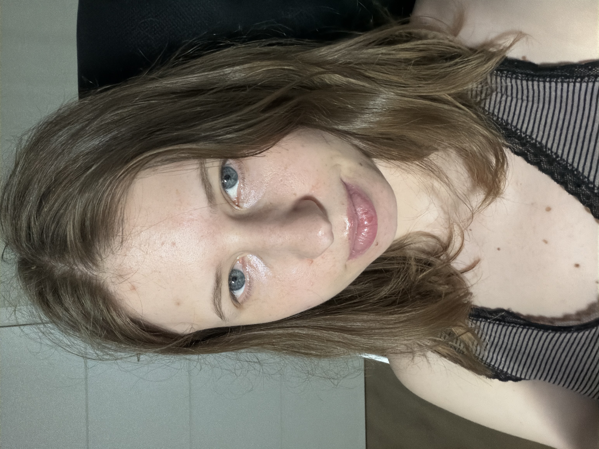 The image is a close-up, selfie-style photograph of a young woman with light skin, brown hair, and blue eyes. She is wearing a black and gray striped top with lace trim. The background is indistinct, but appears to be an indoor setting with a textured wall and a dark chair. There are no discernible landmarks or contextual clues to indicate a specific location in Dongducheon-si, South Korea, or any other city. The image focuses solely on the woman's face and upper body.