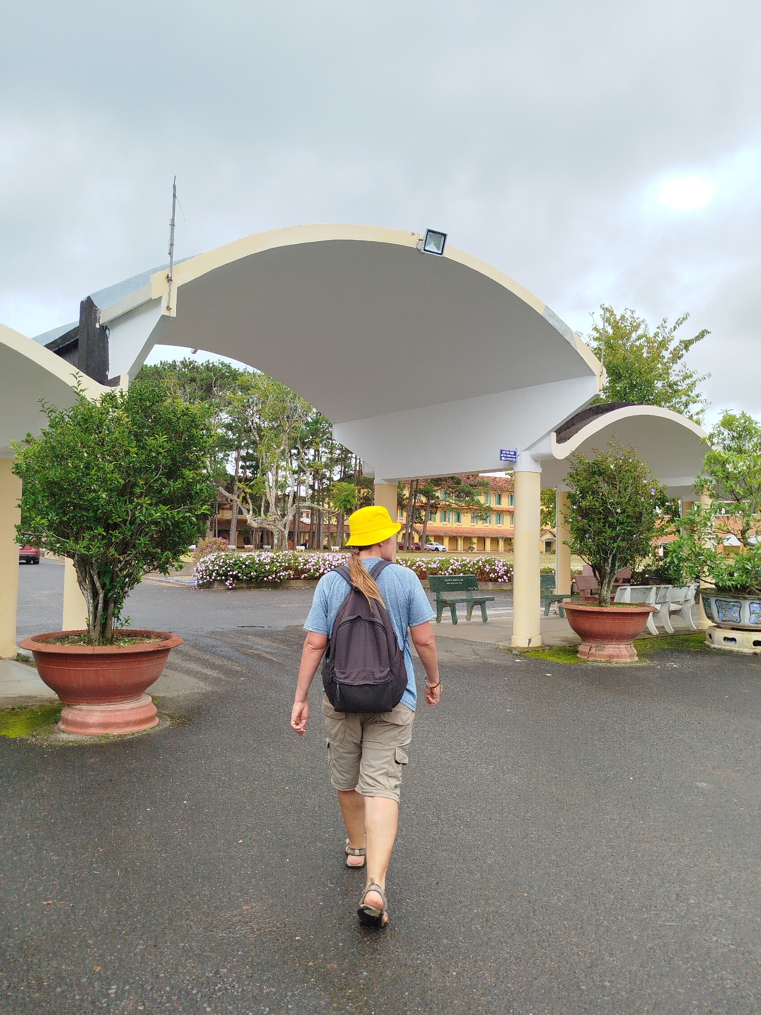 A person wearing a yellow hard hat, blue t-shirt, and tan shorts walks away from the camera toward a covered walkway. The person carries a black backpack. They are walking on a paved surface that appears wet. Large potted plants with green trees are on either side of the walkway. In the background, there is a yellow building with a red-tiled roof and more trees and foliage. The sky is overcast. The location appears to be the Xuân Hương Lake area in Đà Lạt, Vietnam, possibly a park or public space with architectural elements.