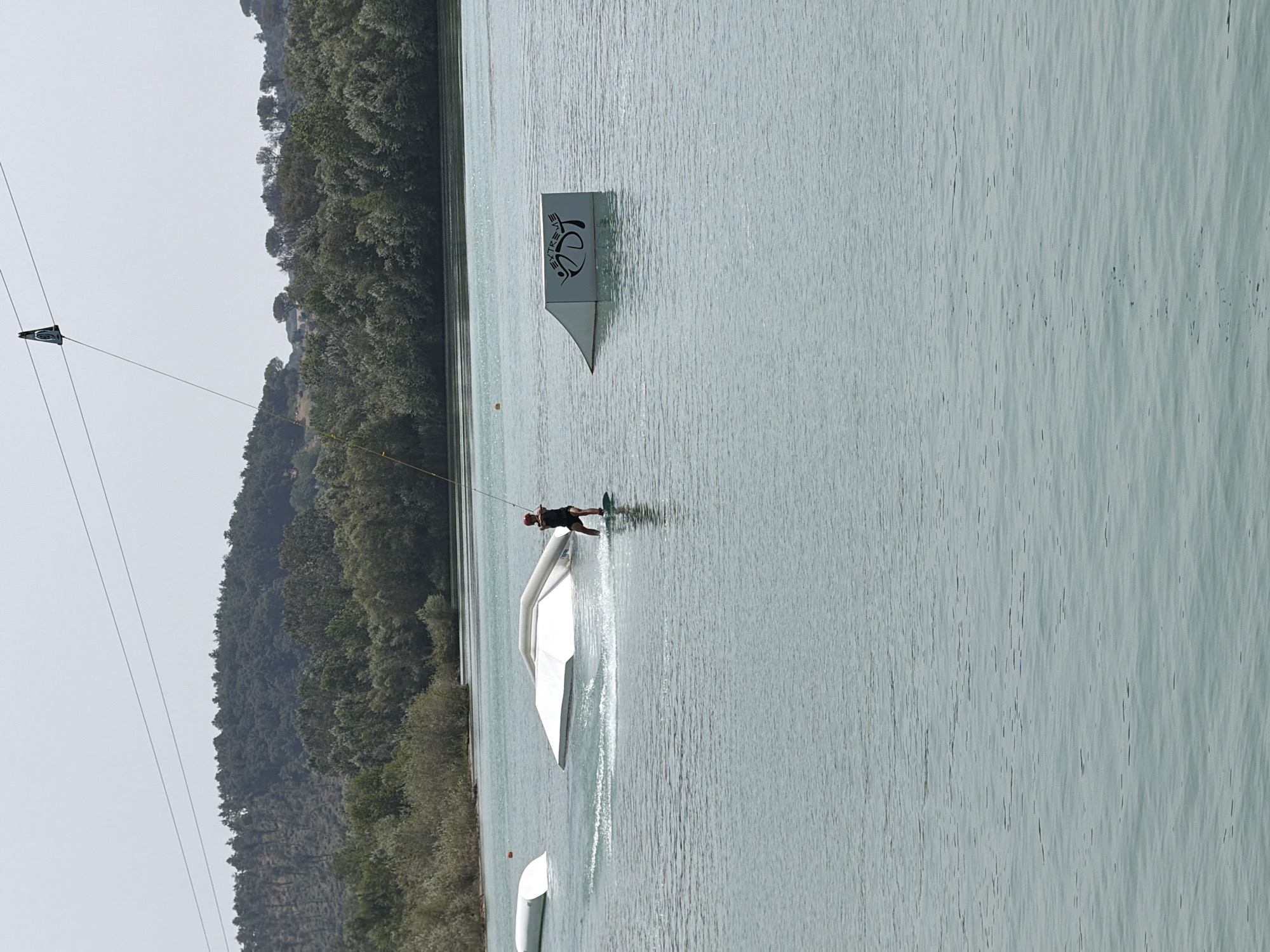 The image depicts a wakeboarding park located on a body of water. In the foreground, a person on a wakeboard is in motion, creating a spray of water. They are being pulled by a cable system that is suspended from overhead wires, with a triangular apparatus visible at the top of the wires. In the middle ground, there are several white obstacles on the water, including a ramp-like structure and a curved, partially submerged object. The background shows a dense line of trees and hills under a hazy sky. The water is calm with a light ripple effect. There is no visible indication of this location being in Rome, Italy, and the setting appears to be a recreational water park rather than a typical urban city environment.
