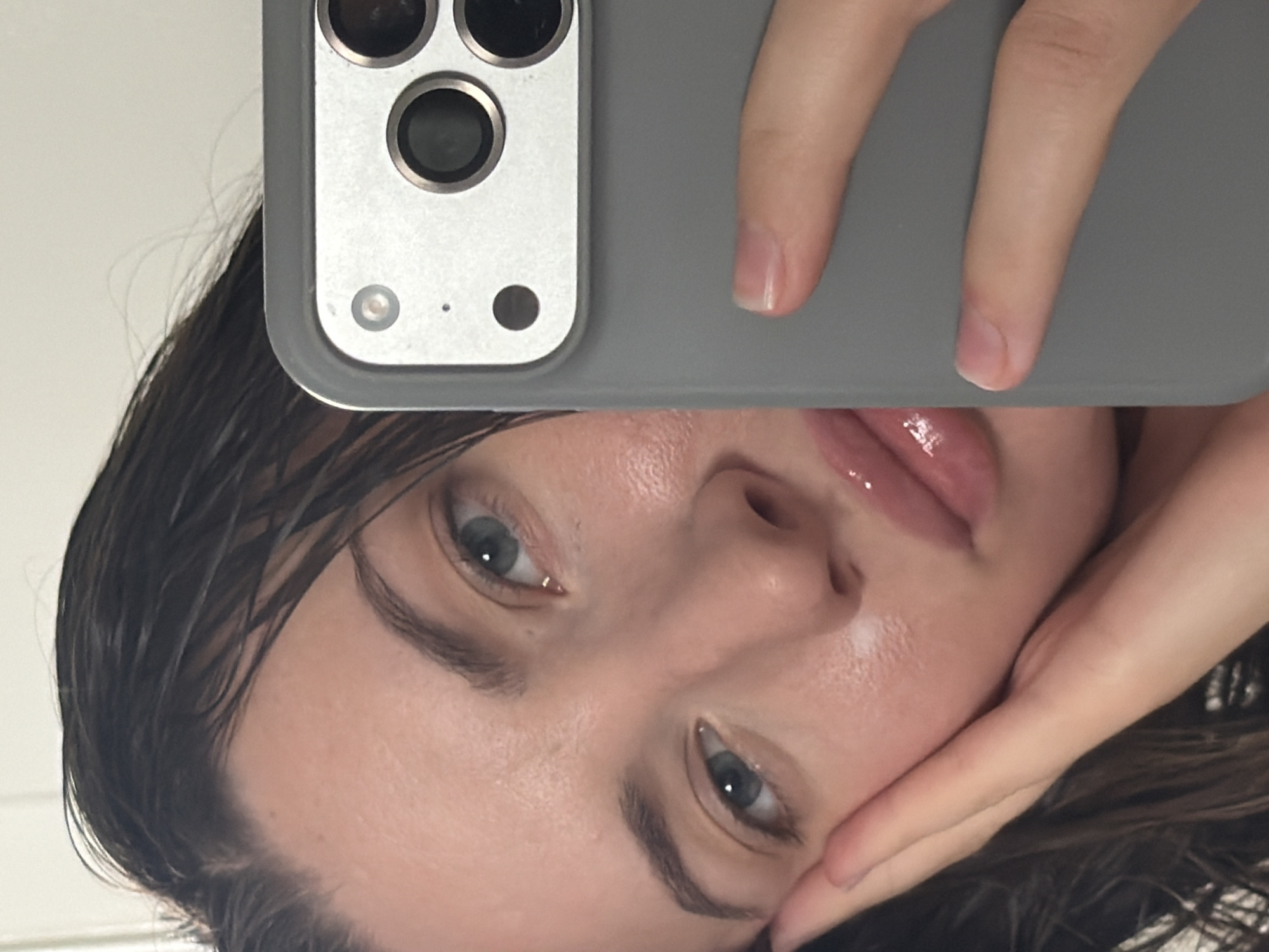The image shows a close-up of a young woman's face as she takes a selfie with a smartphone. Her left hand is placed on her cheek, and her right hand is holding the phone, which is partially visible on the right side of the frame. The woman has wet, dark hair, light blue eyes, and full lips with a glossy finish. Her skin appears dewy and has a subtle glow. The background is plain and light-colored, suggesting an indoor setting. There are no discernible elements in the image that indicate the location of Sharm El Sheikh, Egypt.