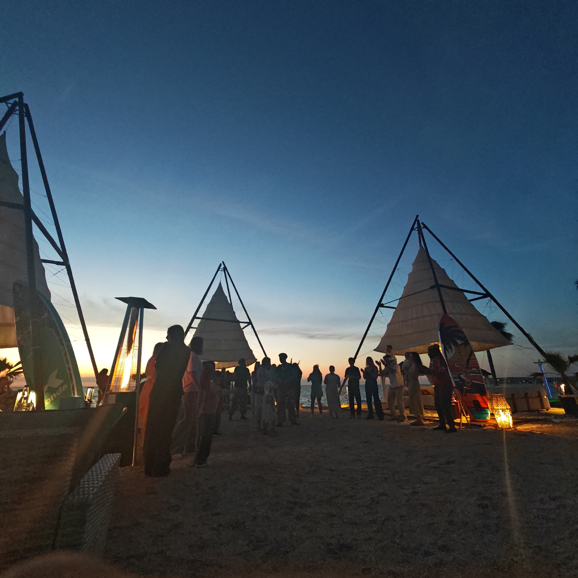 At dusk on a beach in Mysivske settlement, Ukraine, a group of people are gathered. Three prominent triangular structures with canvas sails are erected on the sand. In the foreground, a tall, outdoor heater emits a warm glow. Several surfboards are leaning against the structures. People are standing, talking, and some appear to be watching an activity or performance. The sky is a gradient of deep blue and orange, indicating the sun has recently set. The sea is visible in the distance.