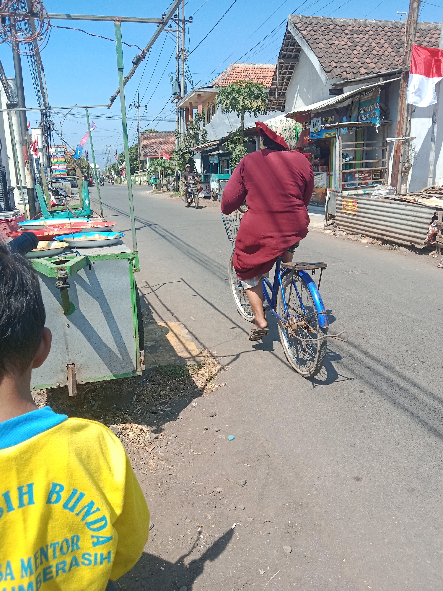 In Mentor, Indonesia, a person is cycling on a street on a sunny day. The cyclist, wearing a maroon dress and a patterned headscarf, is riding a blue bicycle. They are moving away from the camera, towards a row of buildings and shops. A cart with colorful trays is parked on the left side of the street. In the foreground, the back of a person wearing a yellow shirt with blue lettering is visible. The street is paved and there are power lines and electrical poles overhead. A red and white Indonesian flag is hanging from a pole on the right.