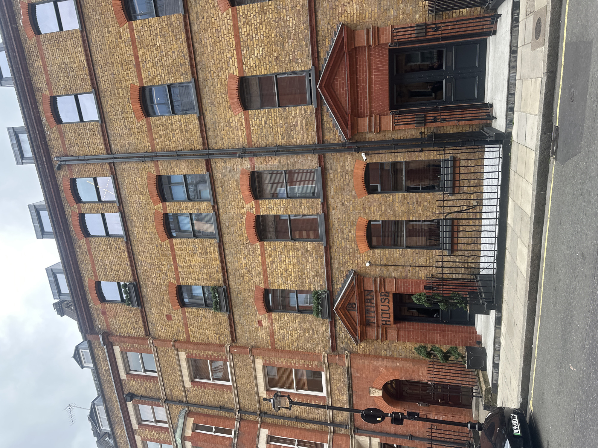 The image shows a brick building in London, United Kingdom. The building, named "Titan House", has a symmetrical facade with multiple windows. The windows on the upper floors have curved brick detailing above them. Black iron railings run along the sidewalk in front of the building. A black car is parked on the street. The sky is overcast.