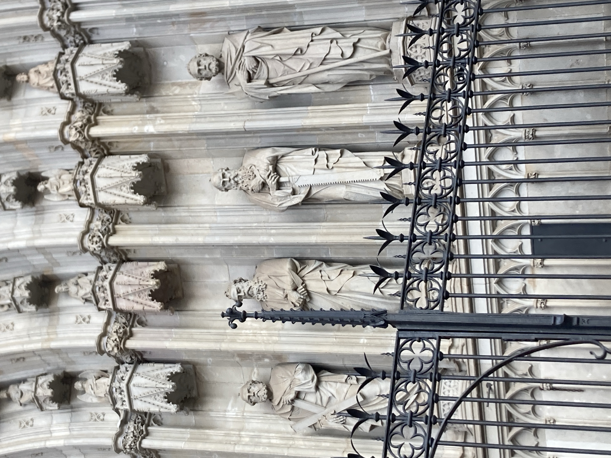 The image shows a close-up view of the facade of the Sagrada Familia in Barcelona, Spain, featuring several stone statues of religious figures. These figures are positioned in niches above a decorative black metal fence. The statues depict bearded men in draped robes, some holding symbolic objects. The architectural style is Gothic Revival, characterized by intricate stonework and pointed arches. The lighting suggests it is daytime, with shadows indicating the sun's position. The overall scene conveys a sense of historical and religious significance.