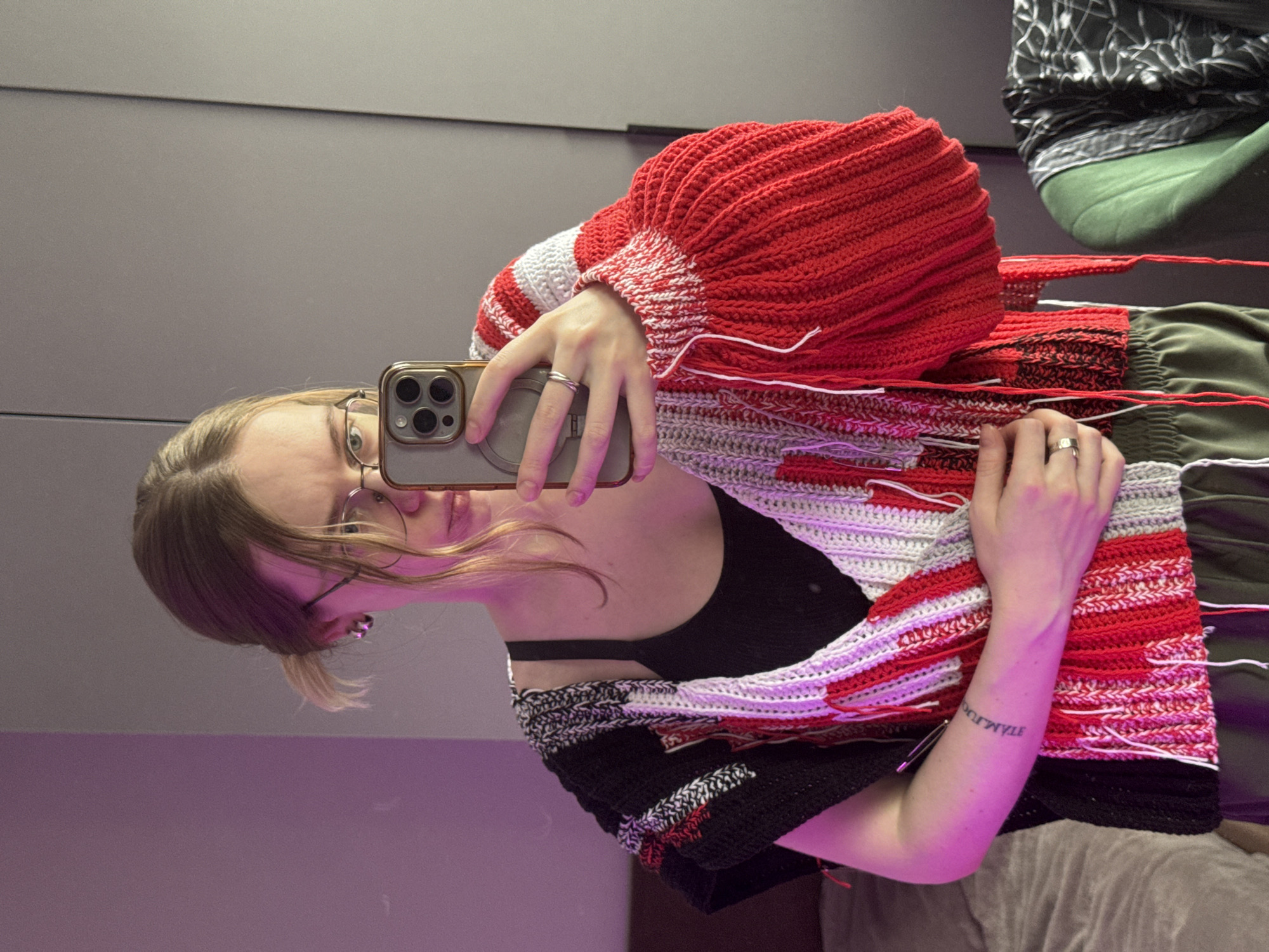 The image depicts a young individual taking a mirror selfie indoors in Krasnoyarsk, Russia. The person, with light brown hair and glasses, holds a gold-toned smartphone, reflecting their face, which shows a neutral to slightly smiling expression. They are wearing a distinctive, possibly handmade, knitted or crocheted garment. This garment features prominent red, voluminous sleeves and is patterned with stripes in red, white, black, and gray yarns. Several loose threads are visible, suggesting it might be a work in progress or a specific design choice. A black top or bra strap is visible underneath the garment.
The indoor setting includes a plain gray wall in the background. The lower part of the reflection exhibits a purple hue, indicative of artificial lighting, possibly during the evening. Other fabrics are partially visible in the background, including a dark, patterned material and a solid green fabric, likely clothing or soft furnishings.
Notable details include a silver ring on one of the person's fingers. On their right forearm, a mirrored tattoo with capital letters is visible, appearing as "STAMJATS" or a similar reversed word, difficult to fully decipher due to the reflection.