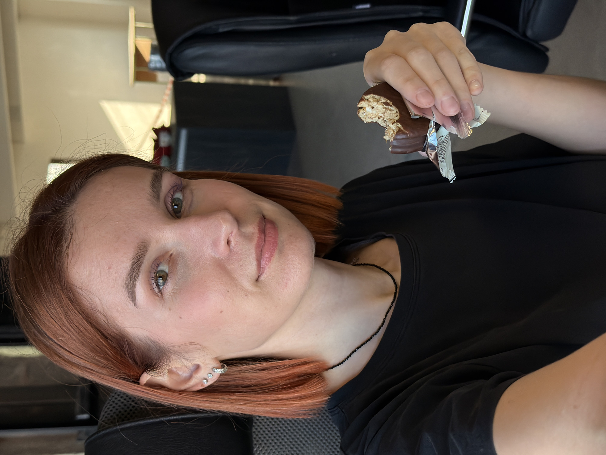 A person with reddish-brown hair and fair skin is holding a partially eaten chocolate bar in their right hand. They are wearing a black shirt and a delicate black beaded necklace. A silver hoop earring and two additional small stud earrings are visible in their left ear. The person is seated in a black chair. The background includes furniture, suggesting an indoor setting, possibly a home or office. The location, Chaikovsky, Russia, cannot be confirmed from the visual information.