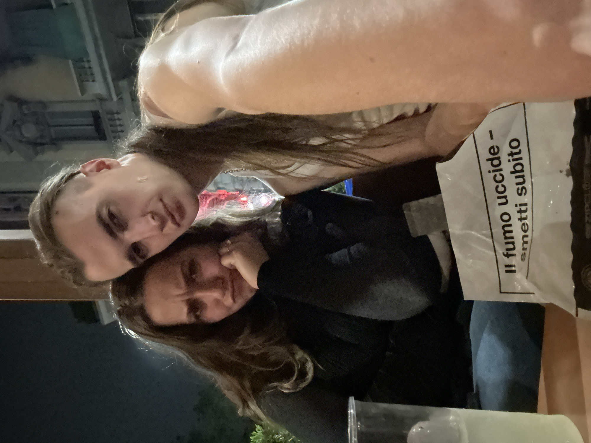 Two light-skinned individuals are posing for a photo. The first individual is on the left, with dark hair and light eyes. The second individual is directly below the first and has brown hair and brown eyes. Both individuals are indoors, next to a table. A plastic bag with Italian text is in the frame near the upper right. Part of a clear plastic cup with liquid inside is visible in the bottom center. Based on the architectural background visible in the upper left, the photo may be taken in Milan, Italy.