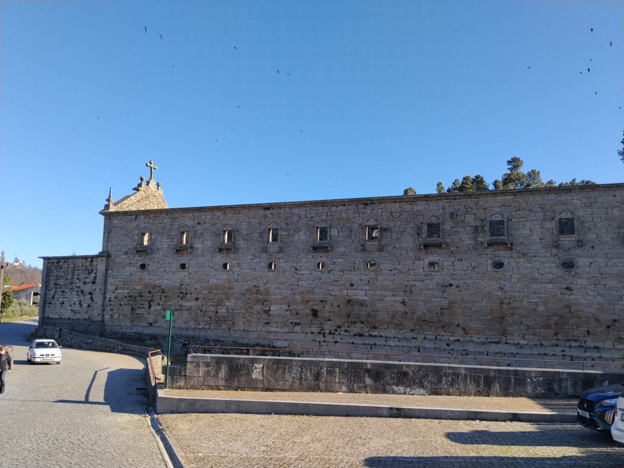 The image depicts the Ucanha Bridge and Tower in Tarouca, Portugal. The stone structure of the bridge and tower features several windows and round openings. A road leads to the bridge, with a white car visible on the road. The area around the bridge appears to be a paved or cobblestone surface. In the background, trees and parts of buildings are seen. The sky is clear and blue. Several small flying birds are present in the sky. There are several cars parked in the lower right corner. A person is partially visible to the left of the white car.