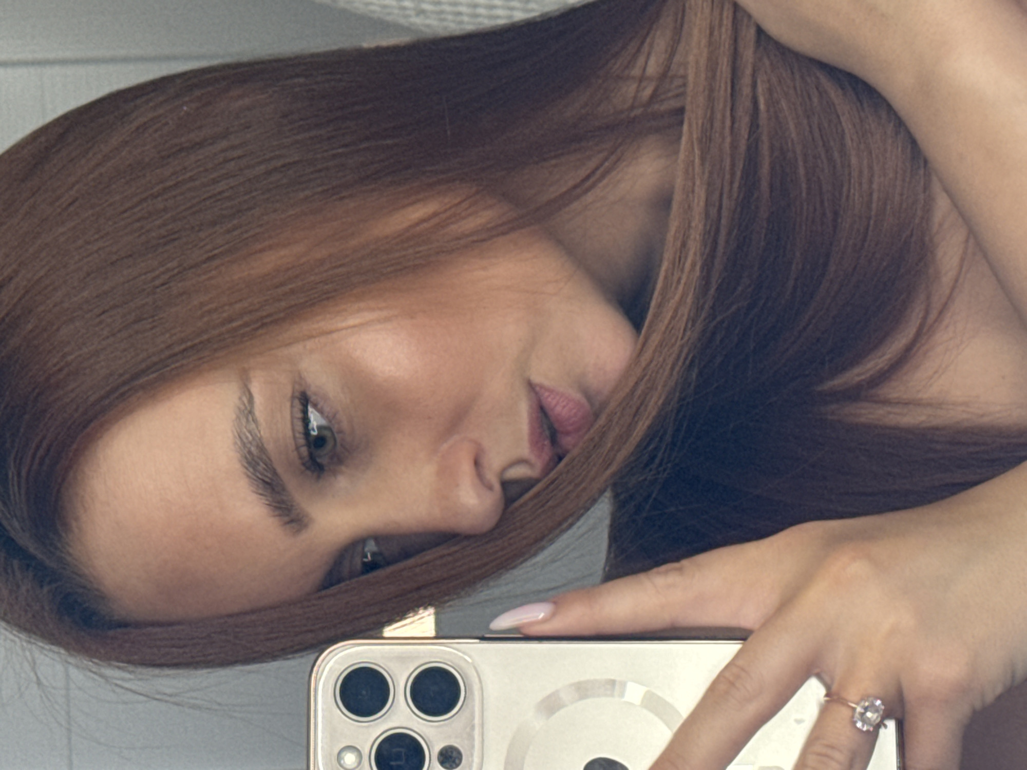 A woman with long, reddish-brown hair is taking a selfie with a smartphone. Her face is partially obscured by her hair and the phone. She is wearing a ring on her ring finger. The location appears to be indoors. There is no information to indicate the location is Chaikovsky, Russia.
