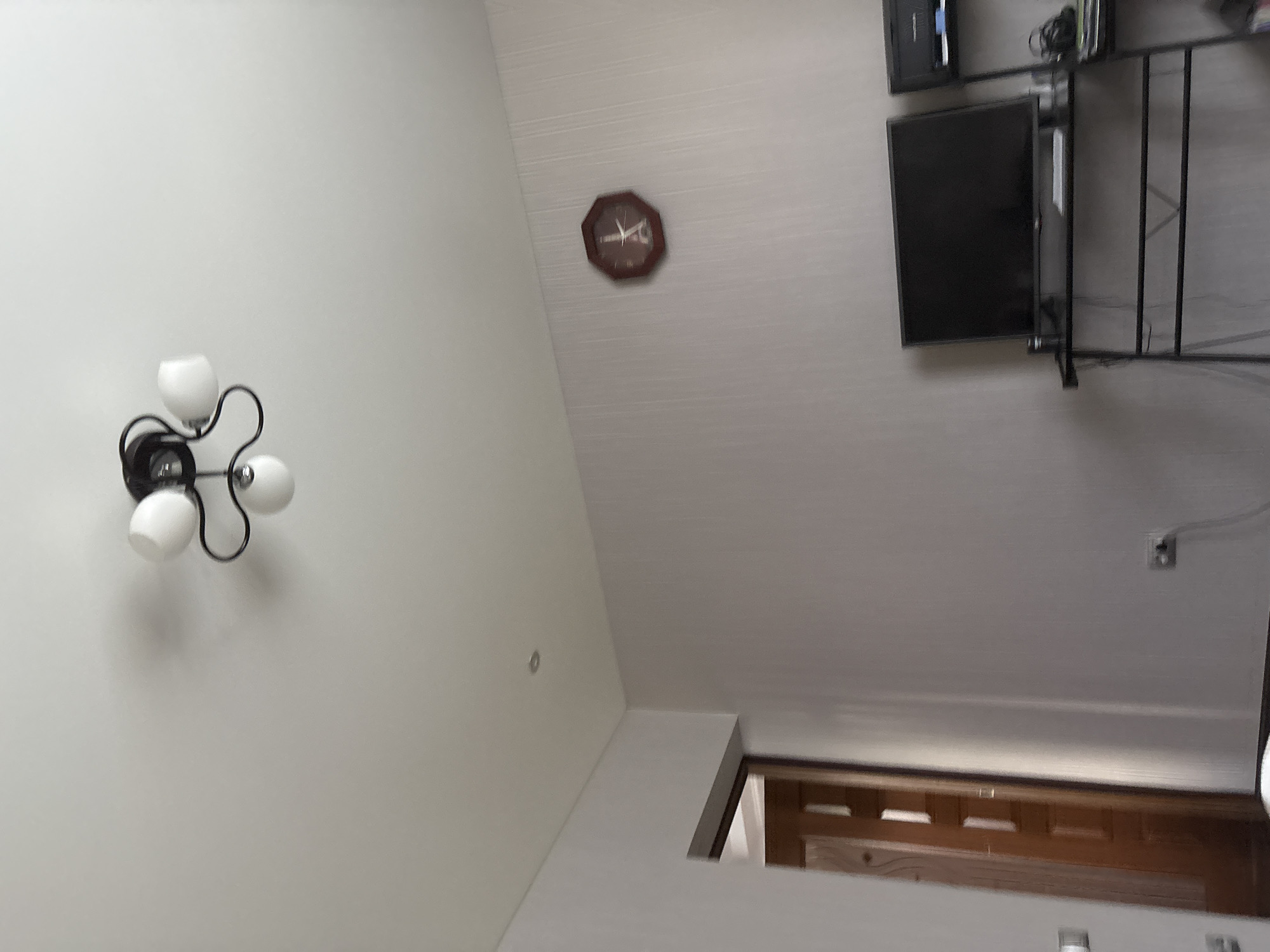 The image shows an interior scene, likely a room in a residential building. A ceiling light fixture with three white globes and a black frame is visible at the top of the image. To the right on the wall, a round, brown clock with a decorative design is mounted. Below the clock, a flat-screen television is placed on a black metal stand. The television is off, displaying a black screen. To the right of the television, another shelf unit is partially visible, holding electronic equipment and cables. A doorway leading to another room is seen on the left side of the image. The walls are painted a light color, and a power outlet with a cord is visible near the floor on the right side of the image. The overall impression is of a living space or a study area. The image does not provide any specific information about the city of Kinel'-Cherkasy, Russia, or any individuals present.