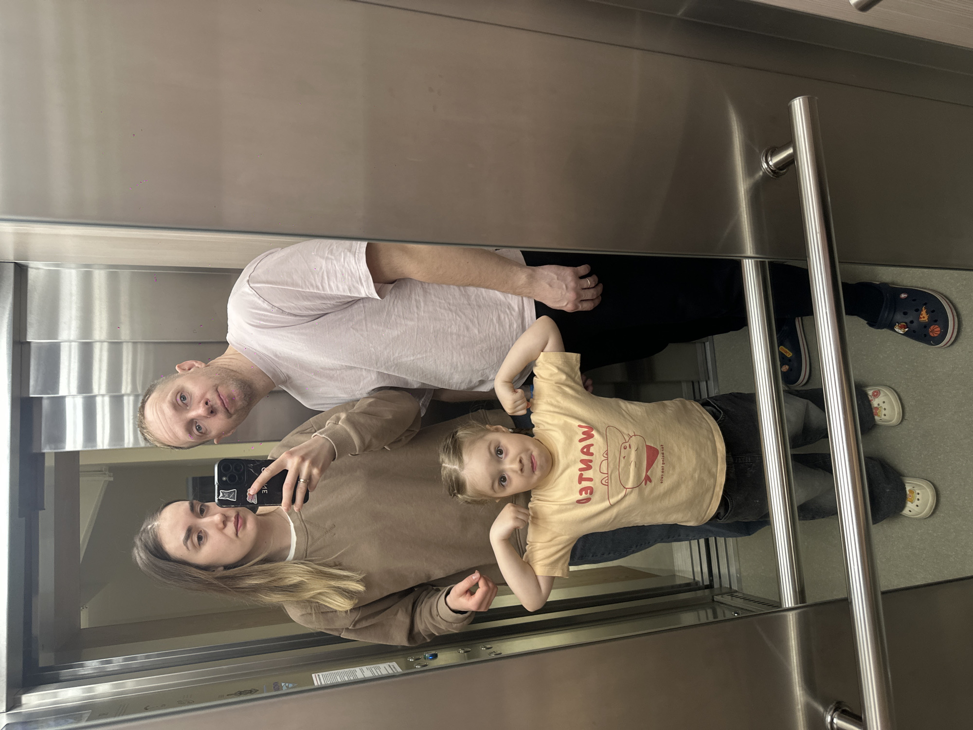 A photograph shows three people reflected in a mirror in what appears to be an elevator. An adult male is wearing a white t-shirt and black pants. An adult female is wearing a brown sweatshirt. A child is wearing a light yellow t-shirt with writing on it and dark-colored pants. The child is posing. The image is taken at an angle. The environment looks like an elevator due to the metal walls, the mirror, and handrails. The location (Glazov, Russia) cannot be determined from the image alone.