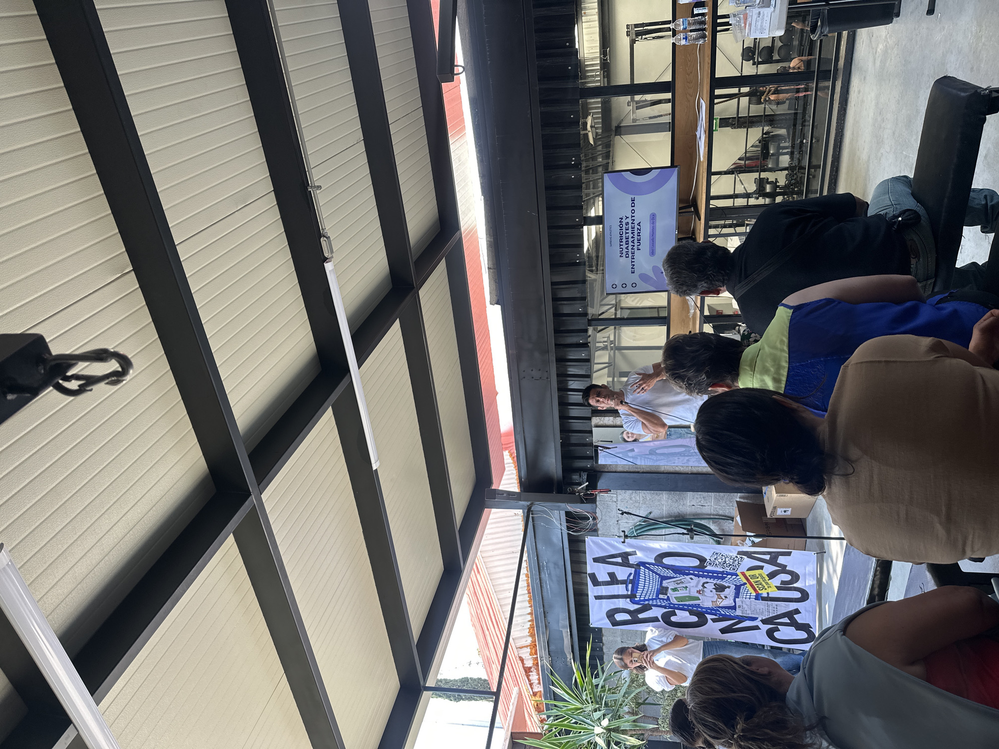 This photograph shows a public presentation taking place at what appears to be an open-air or semi-outdoor venue. The venue has a roof structure with a black frame and what seems to be a corrugated or paneled ceiling. The photo is taken from the audience's perspective.
A speaker is addressing the audience while standing in front of a banner. Another individual is standing to the side, also addressing the audience. The banner reads "RLEA RONA CAUSA," with the word "CAUSA" rotated 90 degrees clockwise. There is an image of a shopping cart on the banner. There is an information stand behind the speaker, as well as a screen to the side, showing the subject matter of the presentation: "NUTRICION, DIABETES Y ENTRENAMIENTO DE FUERZA" ("Nutrition, Diabetes, and Strength Training").
People are seated, facing the front of the room, listening to the presentation. A glimpse through a glass enclosure reveals fitness equipment. A person is visible inside the enclosure. Based on the screen content and the presence of exercise equipment, the event could be related to health, fitness, or a related field.
The OCR text on the screen "Abel Lozada Montes de Oca" is most likely the name of the speaker or presenter. The text on the banner includes the phrase "SCAN OR" which suggests that more information may be obtained by scanning some item (possibly the QR code on the sign).
The location is the city of Puebla, Mexico.