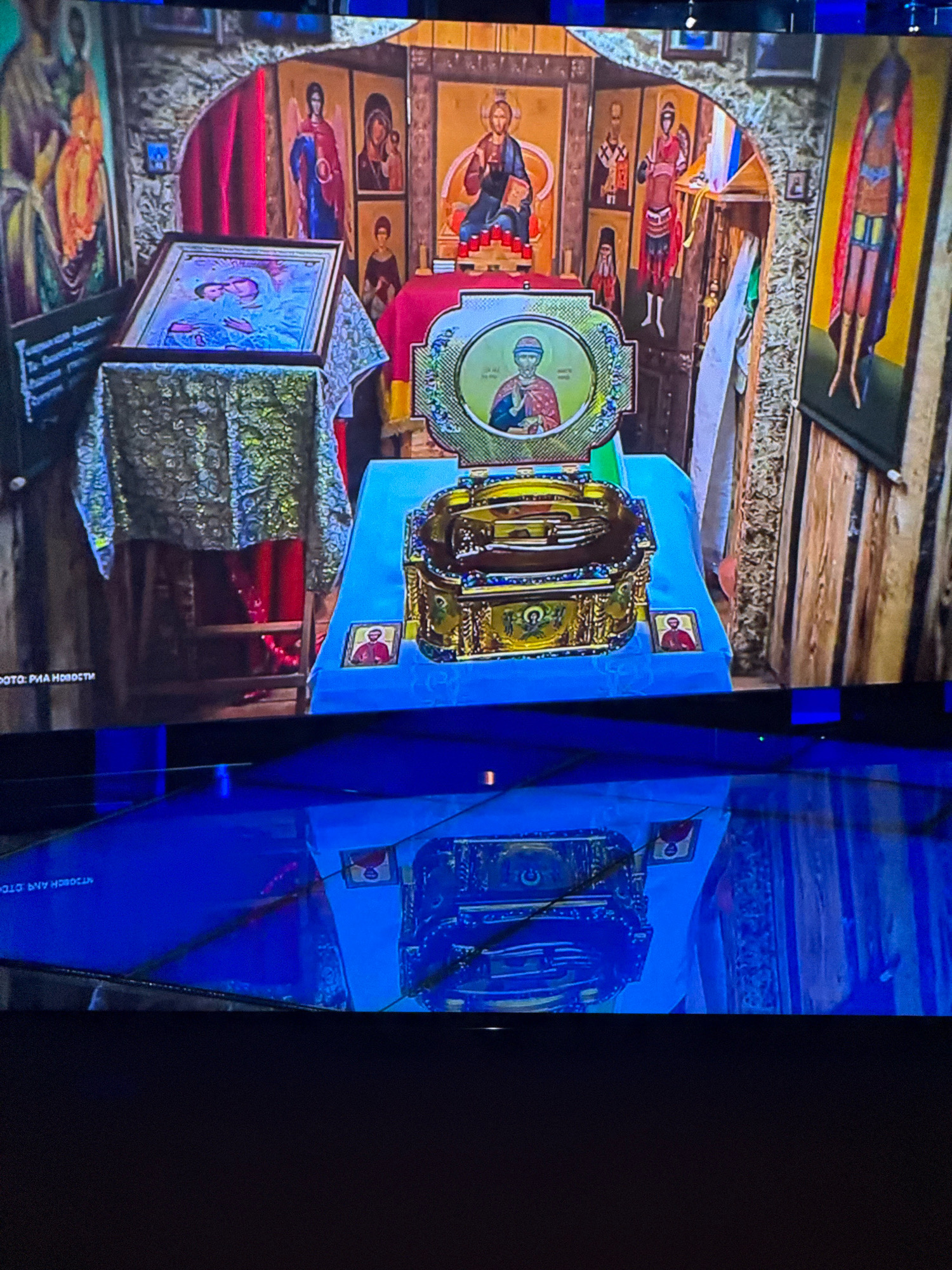 The image captures an indoor scene, likely an Orthodox church or chapel, with a prominent altar area. A framed icon of the Virgin Mary and Child is displayed on a stand covered with an ornate, metallic-patterned cloth. Behind this, various religious icons adorn the walls, depicting figures in traditional attire. At the center is an open, ornate golden reliquary containing what appears to be religious artifacts, possibly bones or fragments, resting on a blue cloth. The environment is richly decorated with religious imagery and symbols. The lower portion of the image shows a reflection of the altar area on a glossy surface, possibly a screen or polished floor. Text at the bottom left corner of the image reads "ФОТО: РИА НОВОСТИ", indicating it's a photograph from RIA Novosti, a Russian state-owned news agency. The location is stated as Khimki, Russia, suggesting the scene is situated within this city.