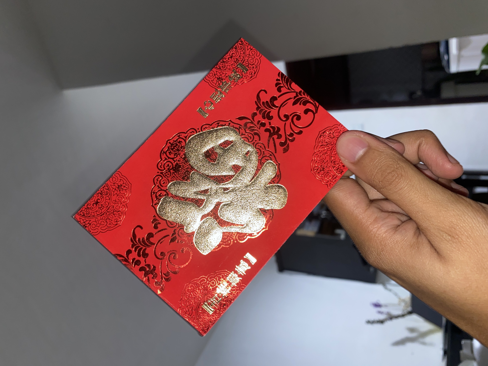 An adult hand, with a medium skin tone, is prominently featured holding a vibrant red envelope (known as an ang pao or hongbao) in a well-lit indoor setting in Davao City, Philippines. The hand holds the envelope by its right edge, with the thumb visible on the front and fingers gripping the back.
The red envelope is rectangular and features intricate gold embossing. A large, stylized Chinese character 福 (fú), meaning "fortune" or "good luck," dominates the center. Surrounding this central character are ornate gold floral and swirl patterns, creating a decorative border and background texture on the envelope.
Two sets of smaller gold Chinese characters are also visible. At the top left corner, rotated diagonally, the characters 恭喜發財 (gōngxǐ fācái), meaning "Congratulations and be prosperous," are embossed. At the bottom right corner, also rotated diagonally, the characters 身體健康 (shēntǐ jiànkāng), meaning "Good health," are visible. The red and gold color scheme, along with the characters, suggests the envelope is used for auspicious occasions, such as Lunar New Year, weddings, or birthdays.
The background behind the envelope is largely out of focus. To the left, a smooth, dark gray surface is visible, possibly a wall or furniture. To the right, behind the hand, there are hints of darker furniture or objects, and a lighter area that might be a wall or a brightly lit surface, with a faint suggestion of a plant. The lighting appears bright and even, indicating either daytime or a well-lit room. The act of holding the envelope suggests it is being presented, received, or admired.