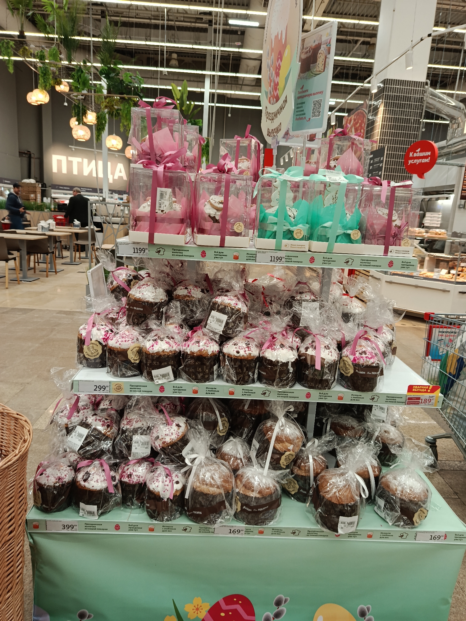 This image captures a festive display of Easter cakes (Kulich) within the bakery section of a large supermarket, likely Auchan, in Moscow, Russia. The scene is brightly lit indoors, with shoppers visible in the blurred background.
The main focus is a multi-tiered stand showcasing an abundance of holiday pastries. The top tiers feature individually packaged cakes in clear plastic boxes, adorned with either pink or teal ribbons, priced at 1199. Below, numerous smaller Kulich are individually wrapped in clear plastic, many topped with white frosting and pink sprinkles, and tied with pink or white ribbons. These are priced at 299.99 and 399.99. The bottom tier displays simpler Kulich in plastic bags, some priced at 169. All products are promoted as "Festive baking from our bakery" (Праздничная выпечка из нашей пекарни) and "Baked with love for you" (Испекли с любовью для вас).
In the distance, a large sign with "ПТИЦА" (Bird/Poultry) is visible, potentially indicating another store section or an eatery. Above the display, promotional signs read "Festive baking" and include a QR code for ordering online at "Auchan.ru". Another sign states "At your service!" (К вашим услугам!). The base of the display is covered with a green cloth featuring Easter egg and pussy willow branch graphics, clearly marking the holiday season. A "BIG DEAL" (УДАРНАЯ ВЫГОДА) sign highlights discounted prices of 189, down from 349.
The image conveys a lively commercial atmosphere, emphasizing readily available traditional Easter treats for customers.