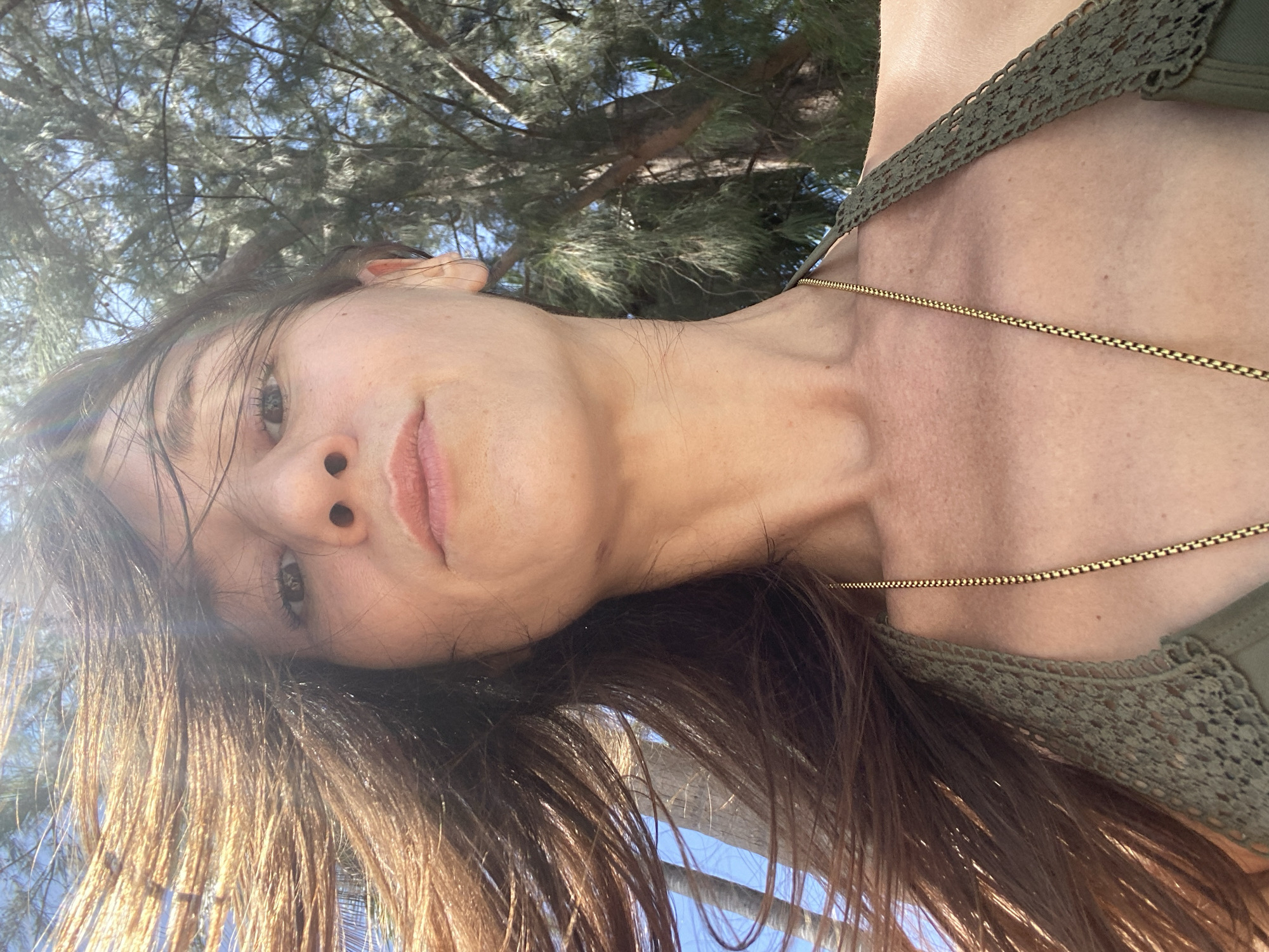 This close-up portrait, oriented at an angle, features a woman looking upwards at the camera. Her face, neck, and upper chest are visible, framed by her long, dark brown hair which is slightly damp and glistens in the bright sunlight. She has a neutral expression, with her lips softly parted and her dark eyes looking directly forward.
She is wearing a dark green top, possibly a swimsuit or camisole, with a distinctive crocheted lace pattern on the strap visible over her right shoulder. A thin gold chain necklace is worn around her neck, resting on her collarbone.
The background is dominated by the dense, green needles and branches of trees, likely casuarina trees, set against a clear, bright blue sky. The strong sunlight and clear sky suggest a warm, sunny day. The upward angle of the photograph, combined with the tree types, indicates an outdoor setting, possibly a beach or park area consistent with Moo 1, Thailand. There is no visible text in the image.
