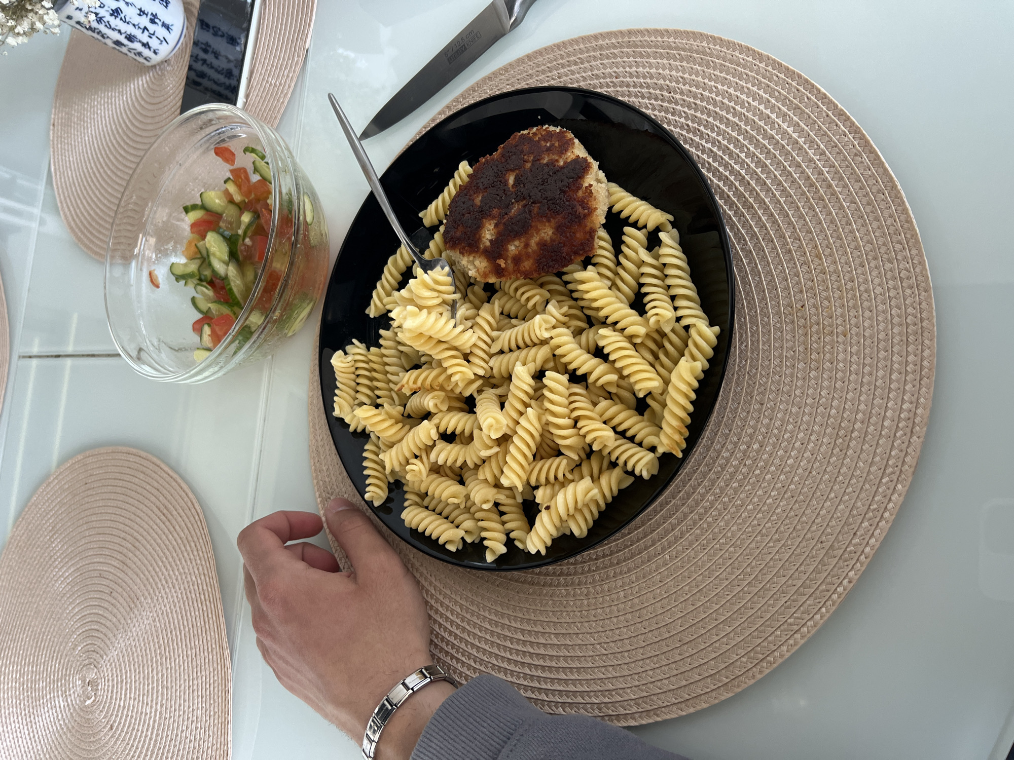 An overhead view captures an indoor dining scene. A white table surface supports a meal presented on two beige, circular woven placemats. Centrally, a black round plate holds a serving of light-colored fusilli pasta and a single, dark-browned, pan-fried patty or cutlet. A silver fork rests on the pasta, partially touching the cutlet. To the right of the plate, a silver-handled knife, bearing the text "KÜCHEN KRAFT," lies on the placemat. A clear glass bowl containing a diced cucumber and tomato salad is positioned above the main plate. To the left, a male-presenting left hand, wearing a silver link bracelet, rests near the edge of the black plate, with a grey garment sleeve visible on the wrist. In the background, behind the salad bowl, a white ceramic vase with blue decorative patterns holds dried white flowers, and a dark mobile phone lies screen-down. The environment appears brightly and uniformly lit.