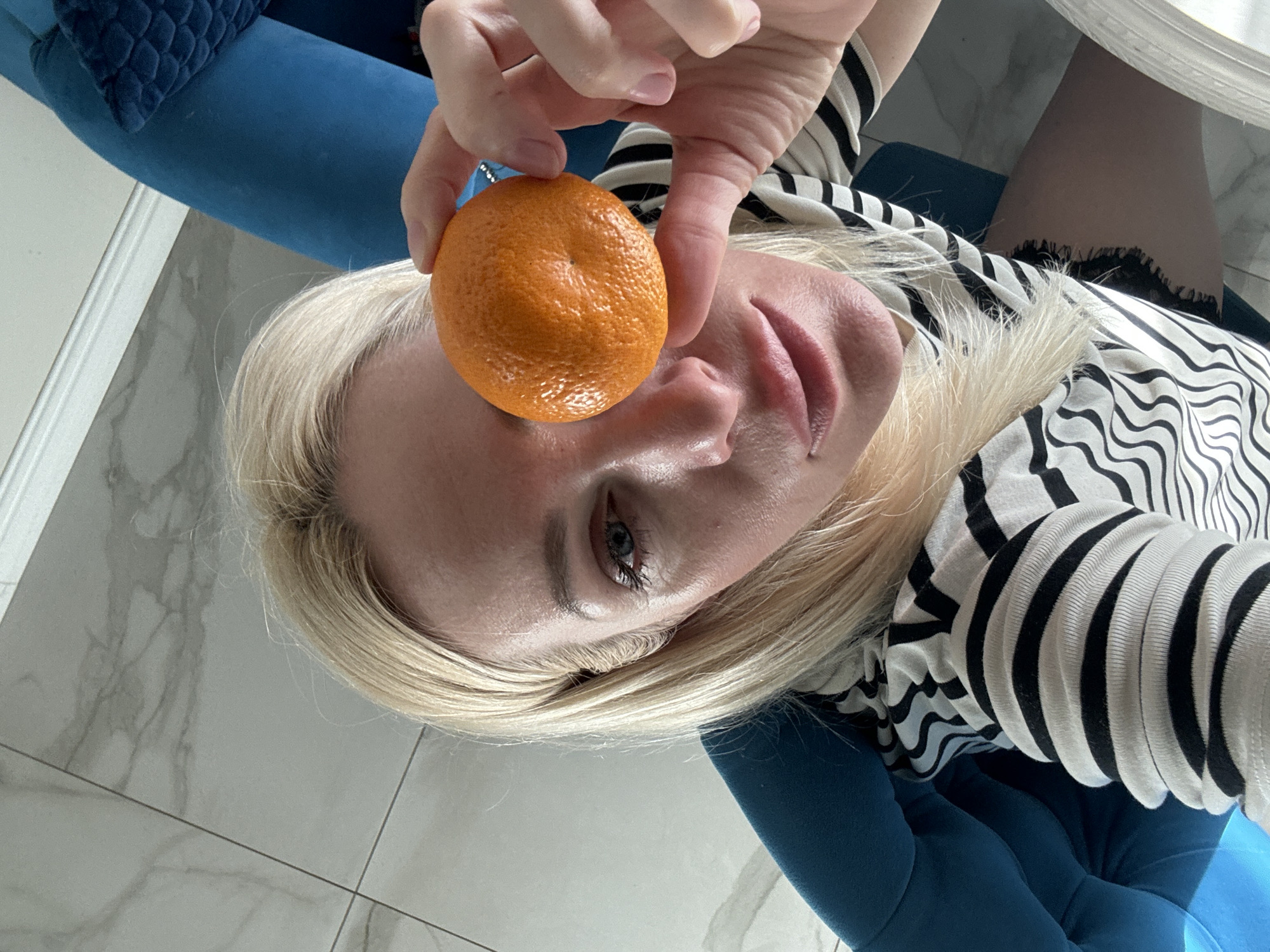 A woman with blonde hair holds an orange in front of her eye, obscuring her left eye. She is wearing a black and white striped shirt and appears to be sitting in a blue chair. The background shows a tiled floor and a white baseboard. The scene is illuminated by natural light.