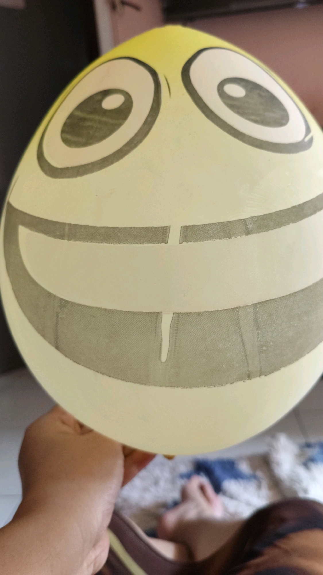 A close-up, slightly angled shot shows a yellow balloon decorated with a cartoonish smiley face. The balloon has two large, circular eyes with pupils and a wide, smiling mouth. The mouth appears to be drawn in two horizontal lines, with a vertical line connecting them, giving the impression of a gap in the teeth. A slight drip of liquid is visible in the center of the mouth. The balloon is held by a person's hand, which is visible in the lower left corner of the image. The background is blurred but appears to be an indoor setting, possibly a living room with furniture and a carpet. The scene is not identifiable as being in Cimahi, Indonesia.