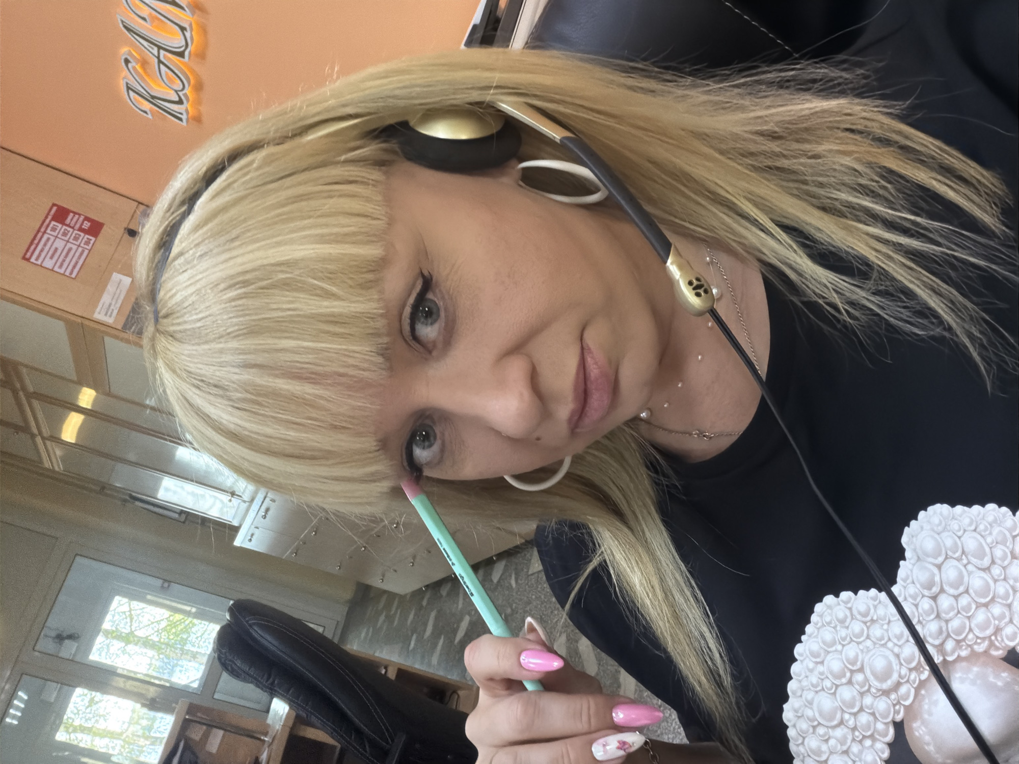 A woman with blonde hair and bangs, wearing a black shirt with a white mushroom design, is applying makeup. She has headphones on and is holding a light blue eyeliner pencil near her left eye. She has pink and white painted nails. The scene appears to be indoors, possibly in an office setting. A window can be seen in the background. There is no verifiable information in the image to indicate this photograph was taken in Chaikovsky, Russia.