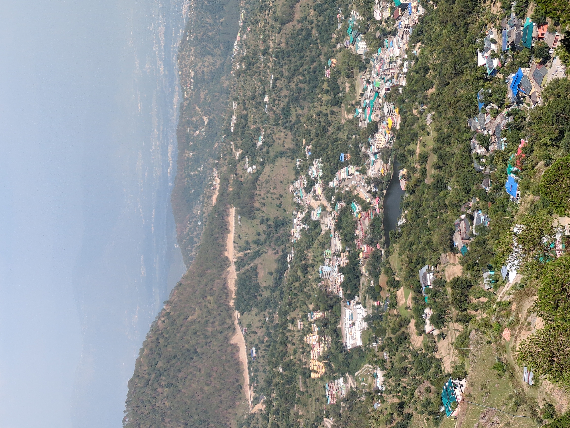 The image presents a panoramic view of a hilly landscape in the city of Doh, India. The foreground is dominated by lush green trees and a steep, rocky incline. As the eye moves further into the scene, the landscape transitions to a densely populated town nestled amidst rolling hills. Buildings of varying sizes and colors, many with distinctive tiled roofs, are scattered across the terrain. A prominent body of water, likely a lake, is visible in the middle ground, reflecting the surrounding environment. A winding road snakes its way up the hillside, connecting different parts of the settlement. In the far distance, a hazy expanse of more hills and a distant urban sprawl suggest the vastness of the region. The overall impression is one of a vibrant community thriving within a natural, mountainous setting.
