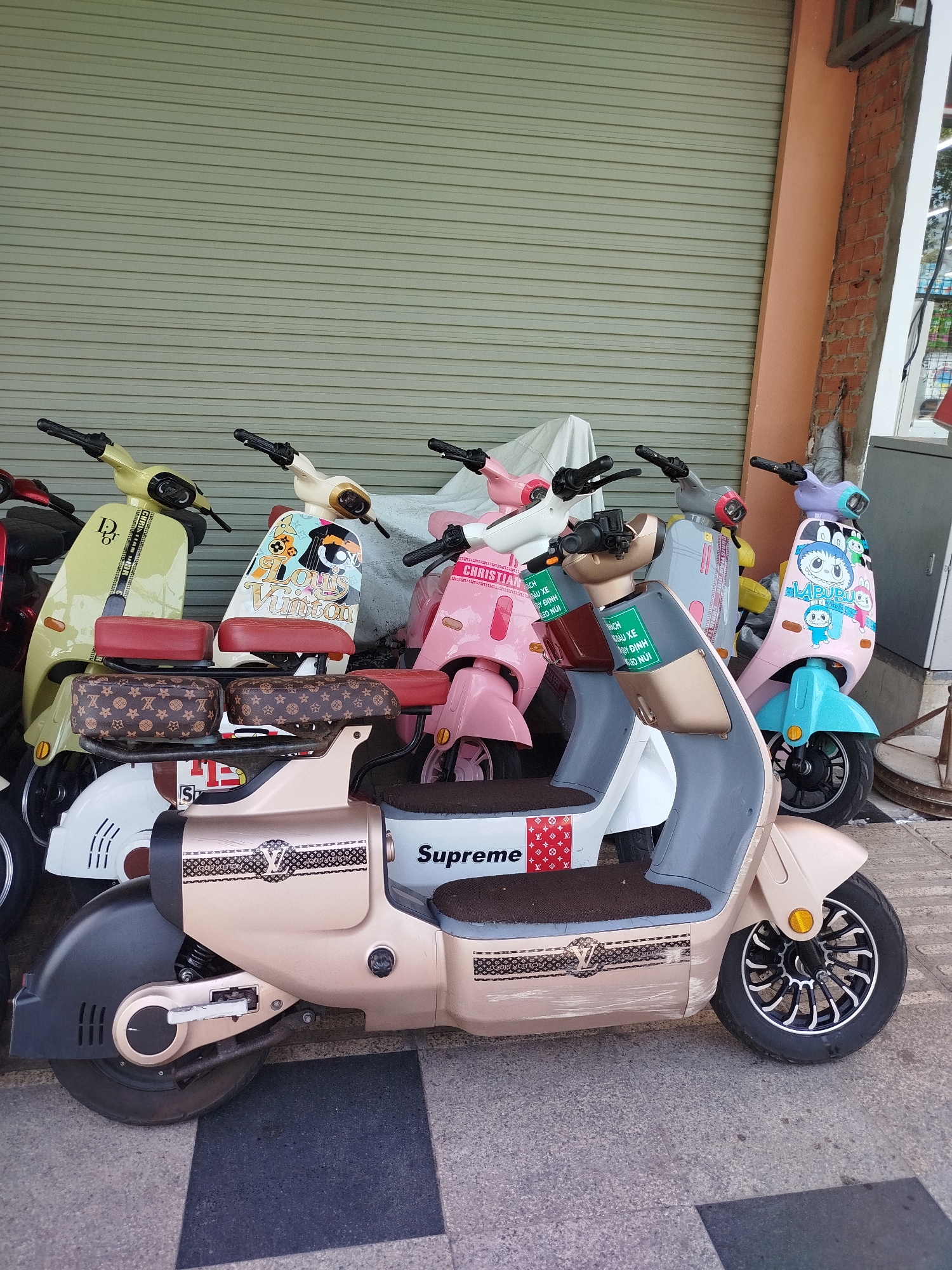 Here's a concise, forensic description of the image:
**Location:** Likely Ho Chi Minh City, Vietnam (based on signage visible).
**Scene:** A row of parked electric scooters, customized with designer branding like "Louis Vuitton," "Dior," and "Supreme," as well as cartoon characters. The scooters are lined up against a closed metal roll-up door and a brick pillar. The scooters are various colors and patterns. Some have Vietnamese signage attached to the handlebars. The ground is paved with gray and black tiles.