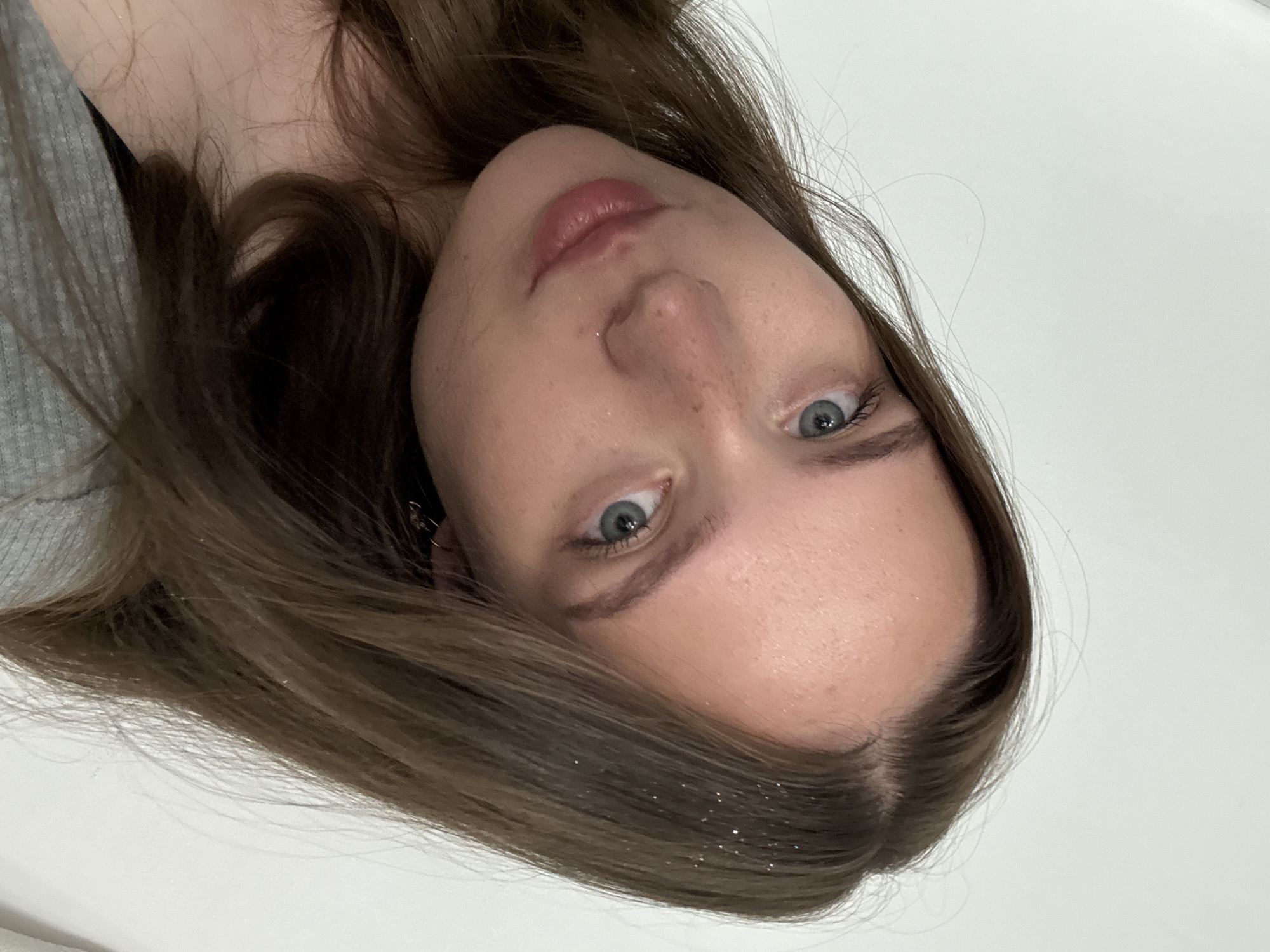 A close-up selfie captures a young Caucasian woman from the chest up, angled to her left, against a plain white background. She has light skin with freckles, blue eyes, and long, light brown hair. Her lips are slightly parted and painted with a soft pink lipstick. Her hair appears to have some glitter mixed in. She is wearing a grey, ribbed garment. The lighting is soft and even, suggesting an indoor setting. There is no visible indication of the city of Chaikovsky, Russia, within the image.