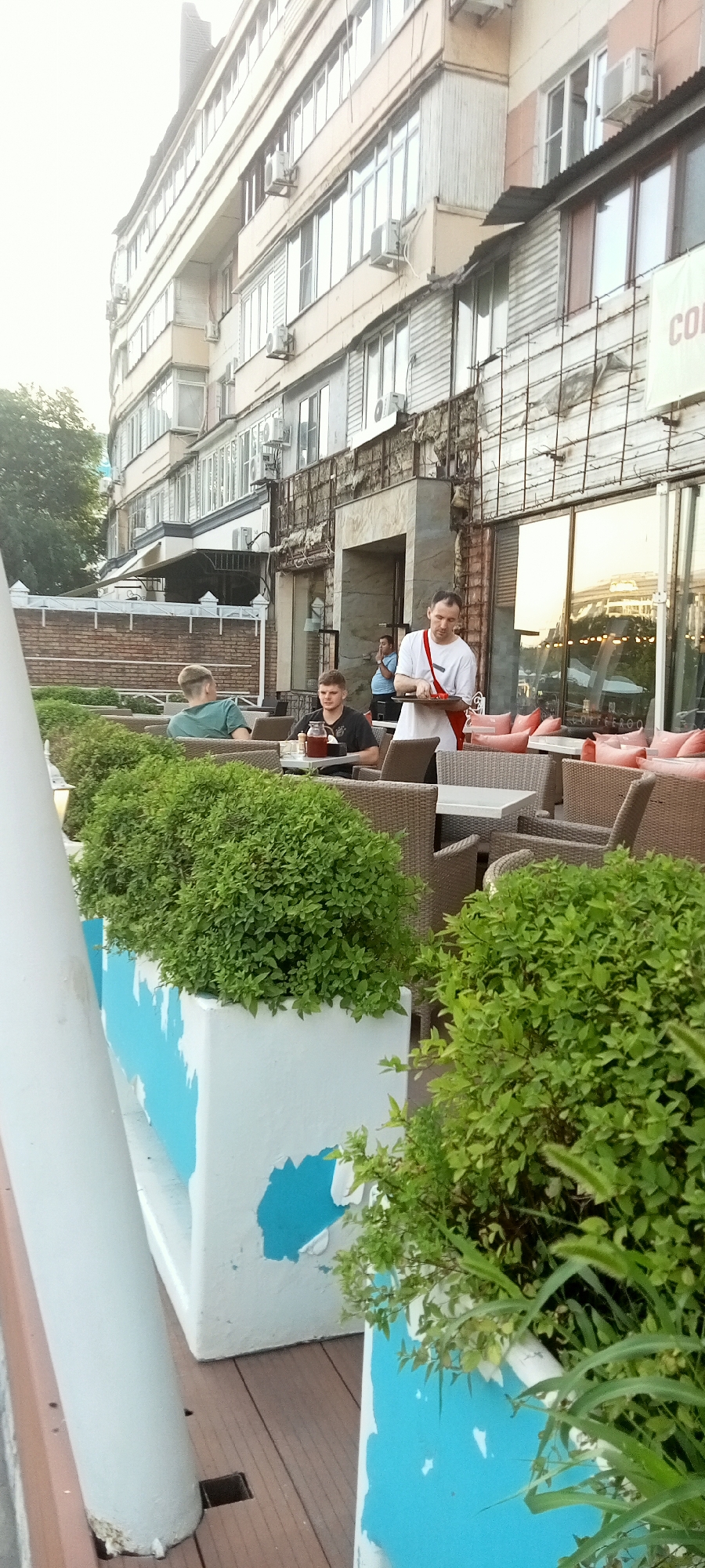 An outdoor seating area of a cafe is depicted in Almaty, Kazakhstan, with a multi-story building in the background. A waiter in a white shirt and red apron is carrying a tray with a drink and a plate of food. Two men are seated at tables, one looking at the camera and the other facing away. Lush green bushes are planted in white and blue planters in the foreground. The cafe has wicker chairs and tables. The building appears to be a mix of old and new architecture, with air conditioning units visible on the facade.