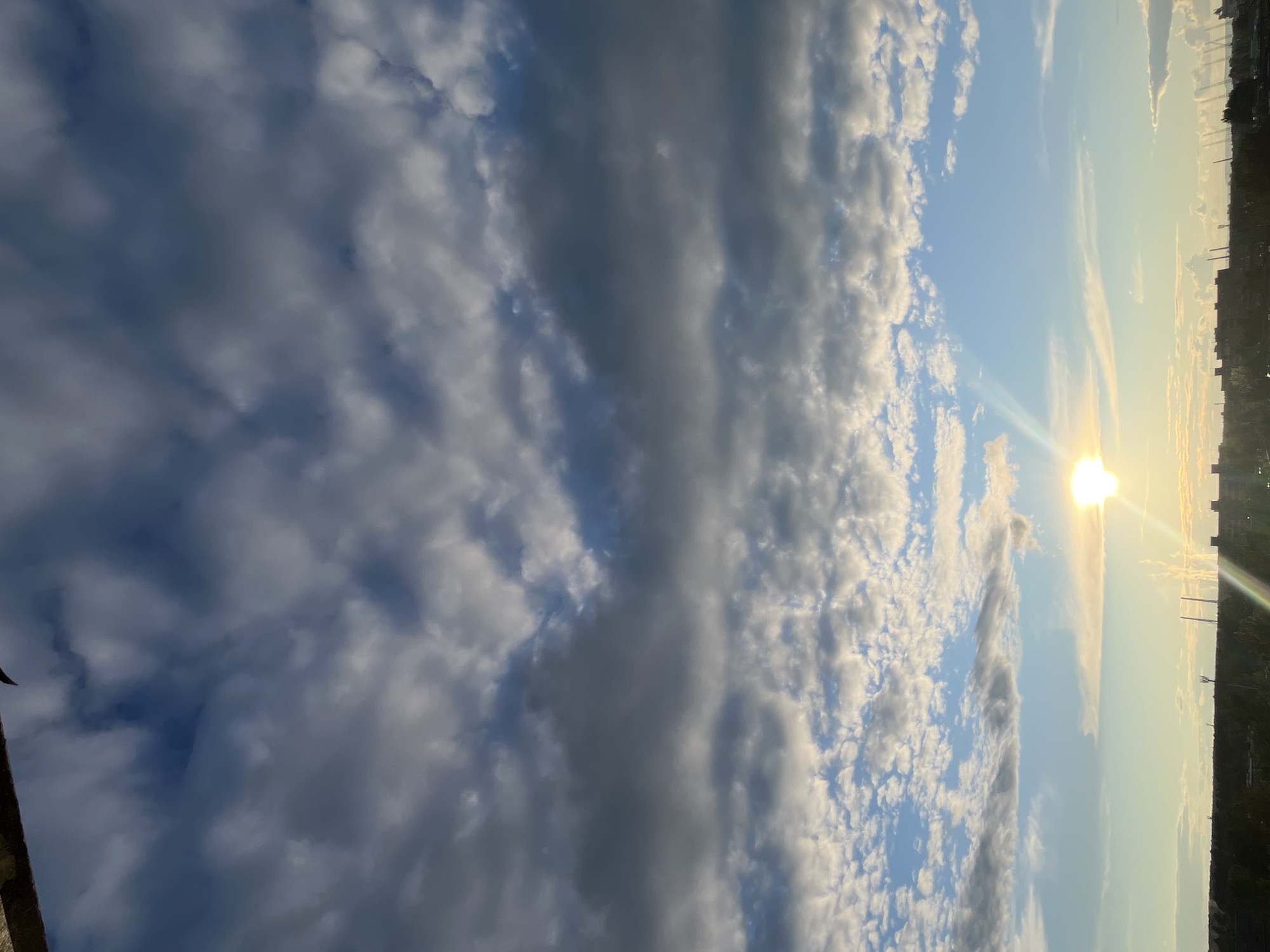 The image captures a daytime sky over an urban area. The sky is predominantly blue, with patches of varying cloud formations. Some clouds are dark and dense, while others are scattered and wispy. The sun is visible, emitting a bright glare. Silhouetted buildings and structures are seen along the horizon, indicating a city skyline. The location is Unknown, Unknown.
