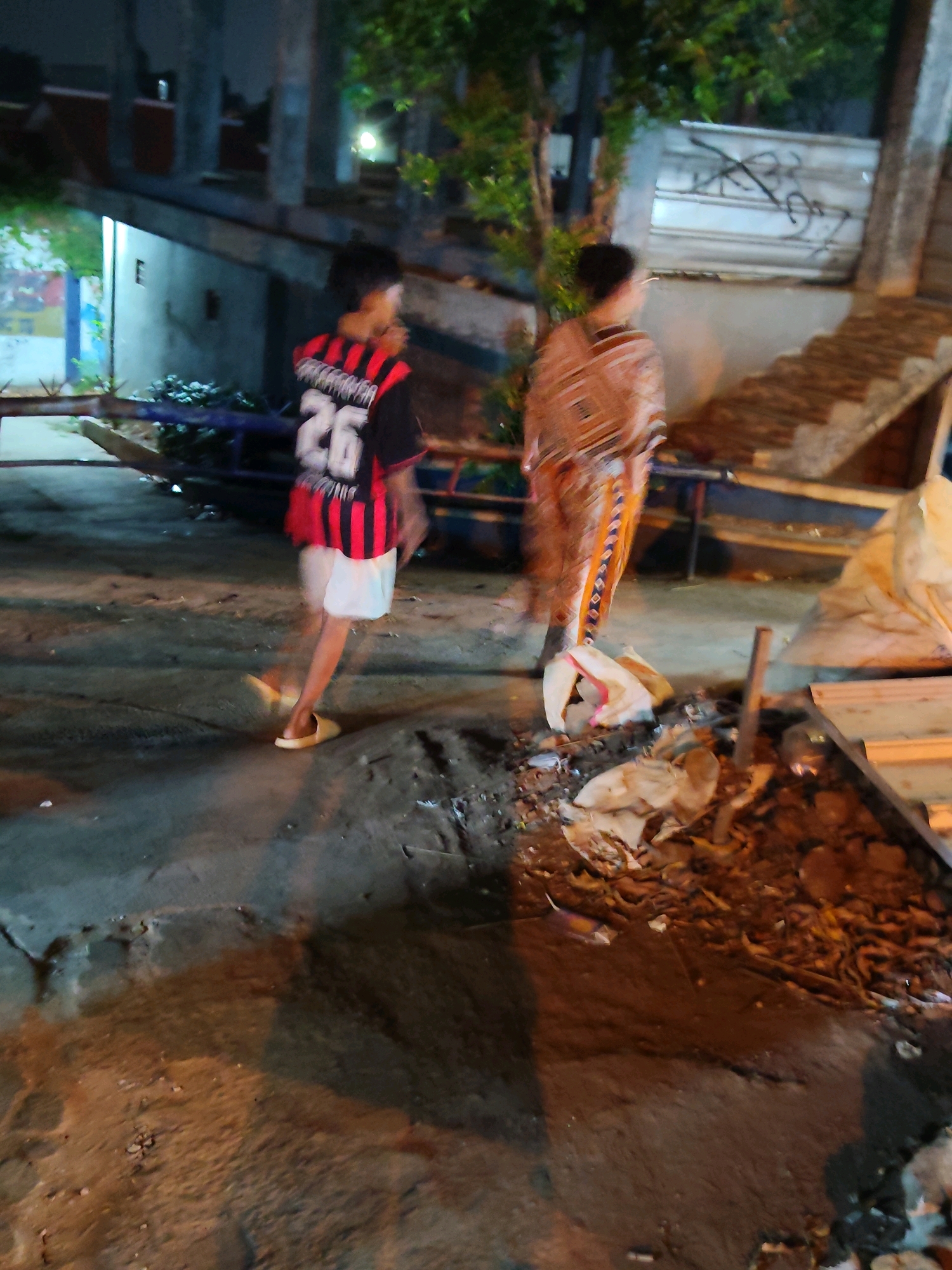 An outdoor, nighttime scene in Purwakarta, Indonesia, depicts two individuals standing on a paved area. The person on the left wears a black and red striped shirt with the number "26" prominently displayed on the back, and white shorts. They are barefoot or wearing light-colored slip-on shoes. The individual on the right is dressed in a patterned garment with geometric designs, appearing to be a sarong or wrap skirt, and a patterned top. They are both facing away from the camera.
The environment includes a rough, textured paved ground with scattered debris, including what appears to be trash bags and organic material. In the background, there are blurred structures, possibly under construction or disrepair, with visible concrete columns and walls. Vegetation, including trees, is present, and a light source casts shadows. A long, boat-like object is partially visible behind the person on the left, positioned horizontally across the scene. The overall atmosphere is dimly lit and somewhat disheveled.