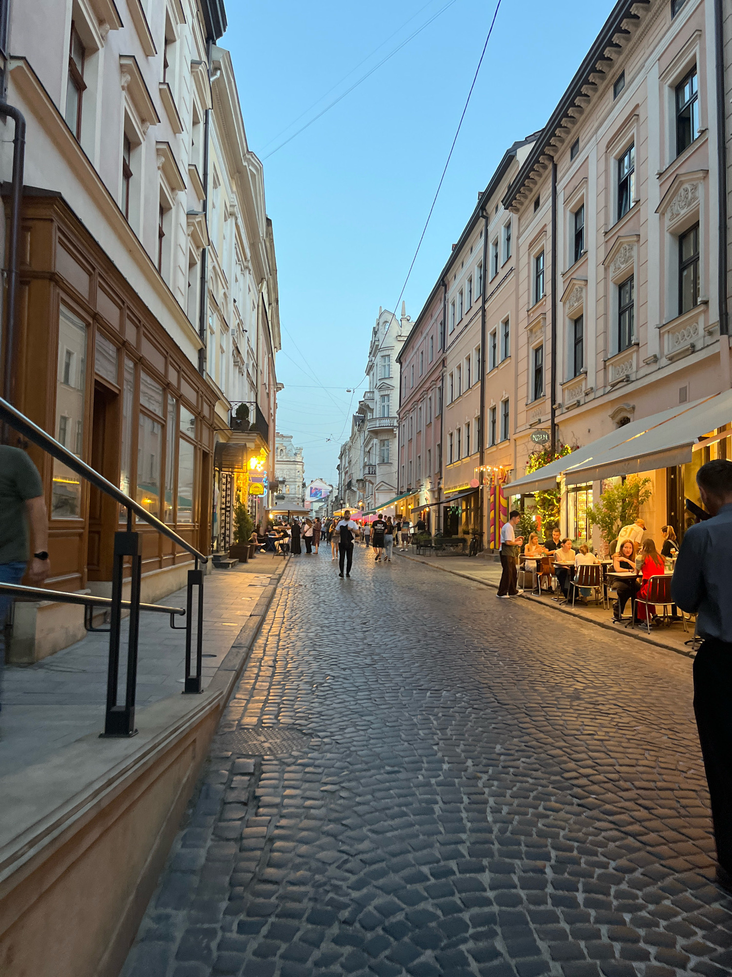 A cobblestone street in Lviv, Ukraine, is lined with buildings featuring ornate architecture and ground-level businesses. Several people are visible, some walking along the street and others seated at outdoor tables of a cafe on the right side of the image. The sky is clear and a pale blue, indicating dusk or early evening. The street is illuminated by streetlights and the warm glow from the cafe, casting long shadows on the cobblestones. The buildings are multi-story with decorative facades and windows. The overall atmosphere is that of a lively, European city street scene.