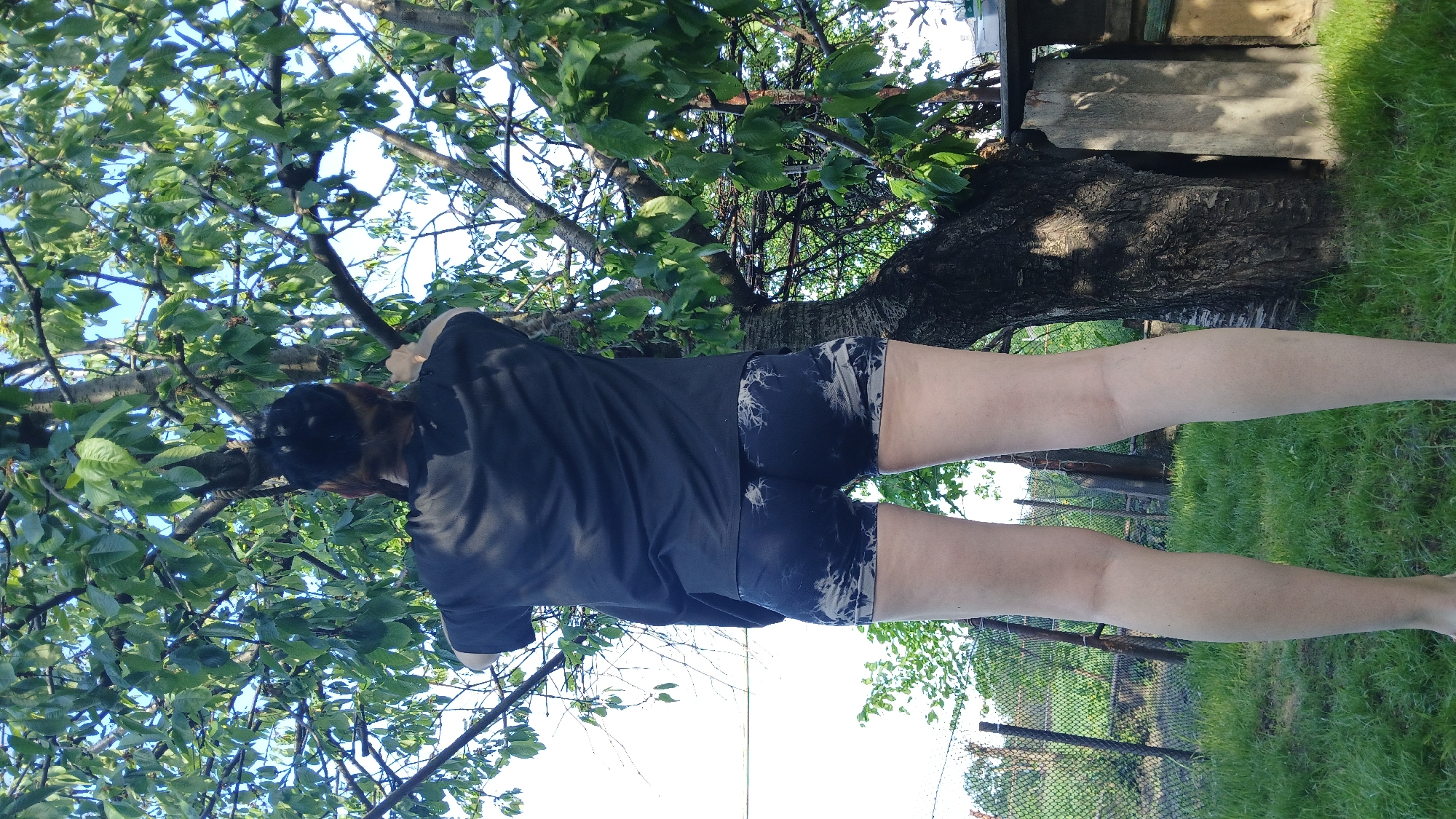 A person is standing under a tree in an outdoor environment. The person is wearing a dark t-shirt and patterned shorts, and their back is to the camera. They are standing on grass, and a tree trunk is behind them. The branches of the tree are overhead, with leaves visible against the sky. A fence and some other greenery are visible in the background. The location is identified as Dnipro, Ukraine.