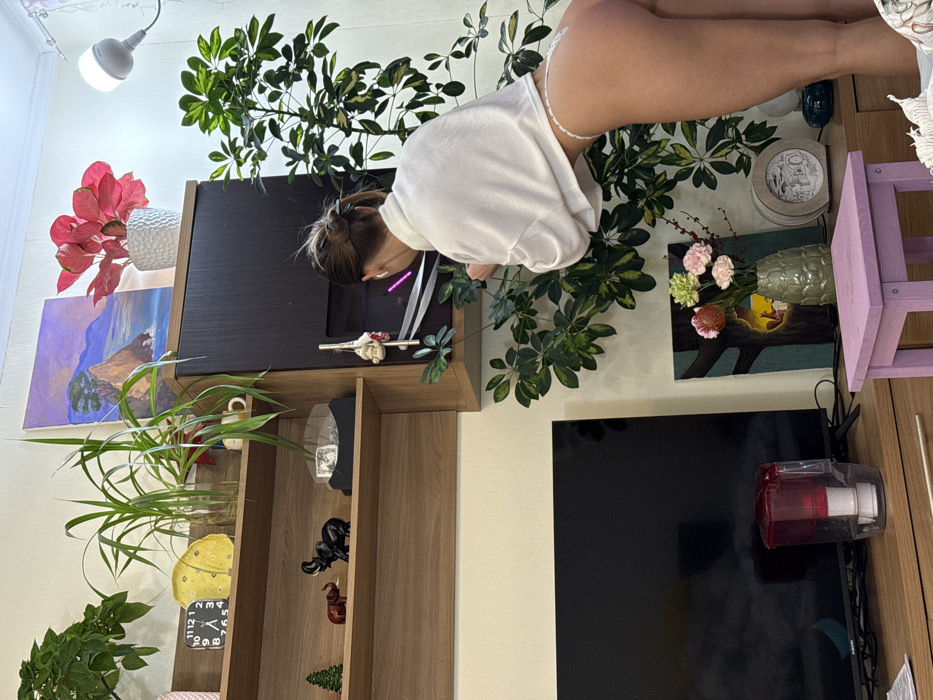 This image depicts an indoor scene within a domestic residence located in Saint Petersburg, Russia.
A single female individual is visible from the back, positioned towards the right of the frame, bent forward and leaning into a large green leafy plant affixed to the wall. She has light brown hair tied in a bun and is wearing a white loose-fitting cropped long-sleeved top, exposing her lower back and buttocks, which are clad in white lace thong underwear. White wireless earbuds are visible in both her ears.
The room features light beige walls and a white ceiling. A light brown wooden shelving unit occupies the upper left portion of the frame, displaying, from left to right: a green potted plant, a black digital clock showing "12:11", a yellow irregular decorative object, a tall green plant in a clear glass vase, and a small plant in a white mug on a lower shelf. Below these, a small green Christmas tree figurine, a reddish-brown elephant figurine, a black elephant figurine, and a clear display case containing a folded banknote (appears to be a US $100 bill) are present.
Above the shelves, a framed painting depicting a coastal landscape with a rocky cliff, sea, and sky is visible. To its right, a red-leafed plant in a white textured pot is displayed. A white ceiling-mounted bulb light fixture illuminates the scene from the upper right corner.
To the right of the shelving unit is a dark brown/black vertical wall unit, featuring a section with a glowing purple light visible through a translucent panel and a small white decorative figure hanging from its edge. The large green leafy plant, possibly a Schefflera, is arranged against the wall and extends downwards, partially obscuring the lower right wall area.
At the bottom of the frame, a light brown wooden console table holds a large black Xiaomi flat-screen television and a red-topped water filter pitcher. To the right of the television, on the console, are a framed artwork depicting a tree with an internal light, a green vase containing artificial pink, green, and red flowers with berries, and a round light-colored decorative frame. A small, light purple wooden stool is positioned in the foreground, towards the bottom right.