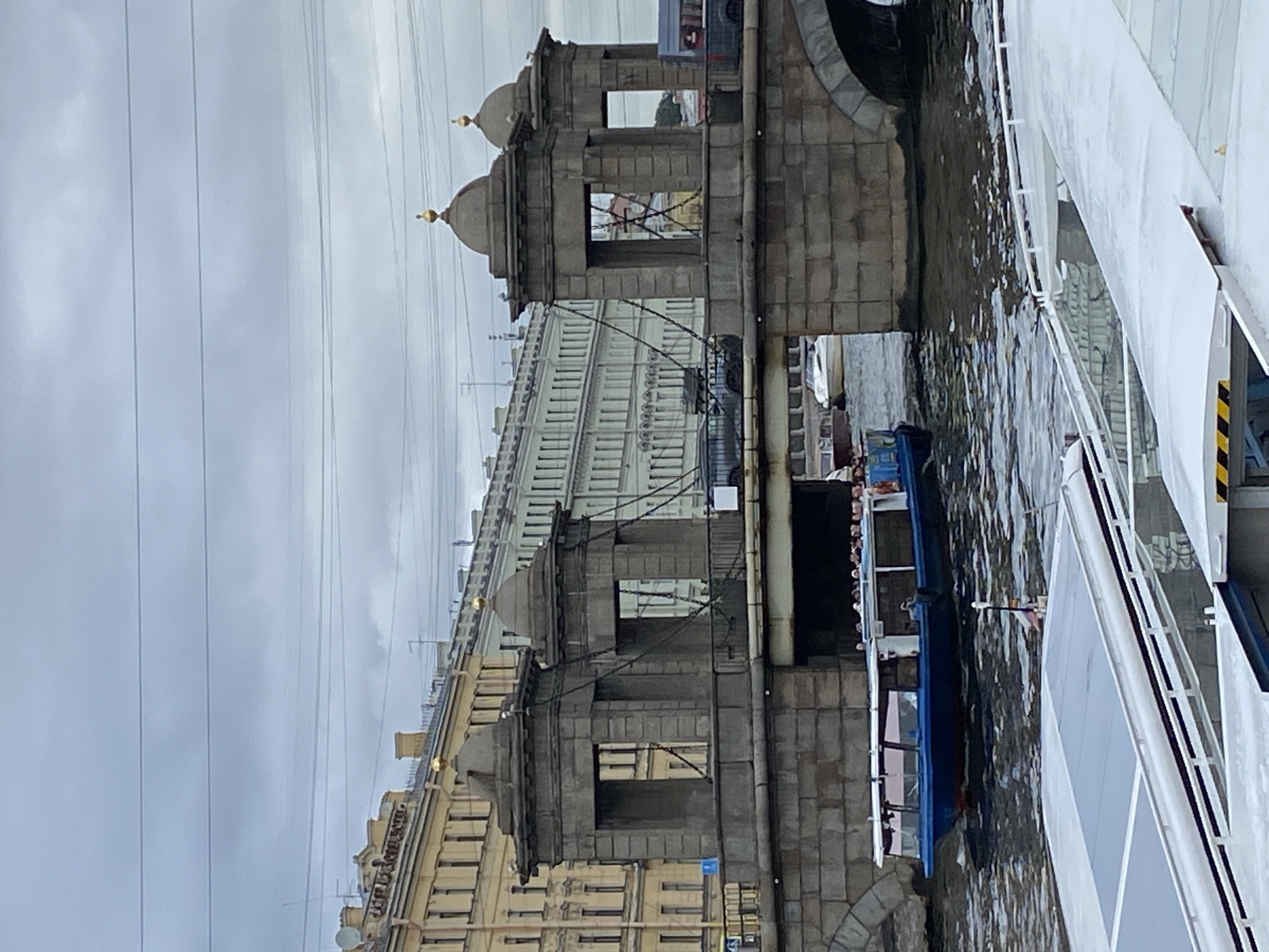 This is a vertical, eye-level, medium shot of a bridge over a canal in Saint Petersburg, Russia, under an overcast sky. The stone bridge has two large, square, stone towers on either side, each with a domed roof topped with a gold finial. Chains hang from the tops of the towers. Behind the bridge, several multi-story buildings with yellow and green facades line the canal. In the foreground, a blue and white tour boat with people on board travels on the dark, rippling water. In the very bottom of the frame, the white roof of another boat is visible. The overall scene suggests a cloudy day in the city.