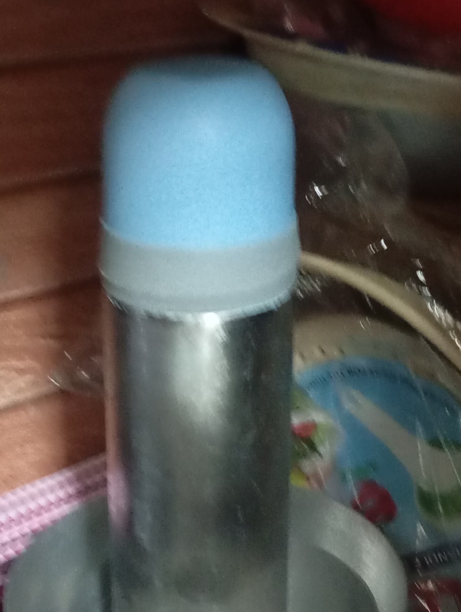 The image shows a close-up of a silver thermos with a blue cap. The thermos is cylindrical and has a textured blue cap on top, separated from the silver body by a translucent white band. The background is blurred, but appears to be a domestic setting with wooden surfaces and possibly other objects. A pink and white checkered cloth is visible in the lower left corner. To the right of the thermos, a circular object with a blue and white pattern is partially visible. The lighting suggests an indoor environment. There are no people or animals in the image.