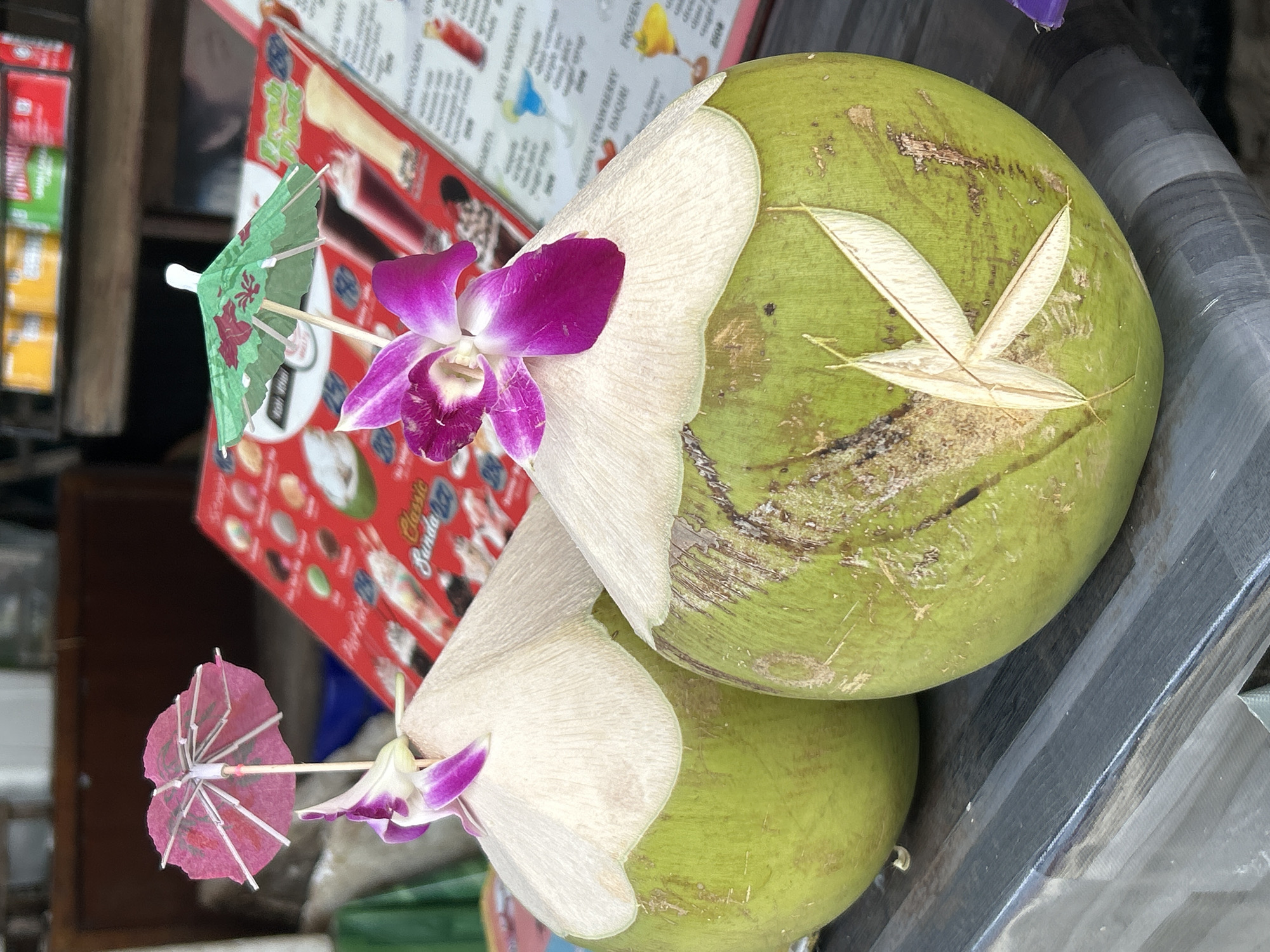 This media file captures a close-up view of two green coconuts, decorated with delicate white petals and vibrant purple orchids. Small, colorful cocktail umbrellas are inserted into the coconuts, suggesting they are prepared for serving. One umbrella is pink with a floral pattern, while the other is green with a similar design. The coconuts are placed on a surface that appears to be a wooden table, with a blurred menu in the background. The menu has images of drinks and text, hinting at a beverage service setting. The overall scene evokes a tropical, possibly beachfront, location. The lighting suggests it is daytime, and the overall mood is refreshing and inviting, typical of a tropical destination like Tambon Phru Nai, Thailand.