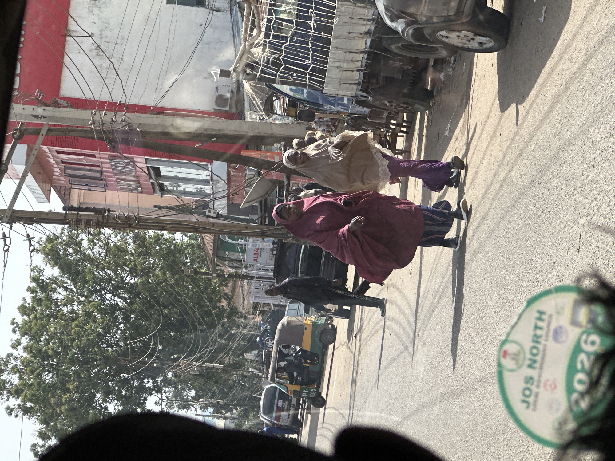 This is a medium shot of a street scene in Jos, Nigeria, taken from inside a vehicle. The foreground shows a blurred green and white sticker on the road with the text "JOS NORTH" and "2026". In the middle ground, two women in hijabs are walking on the right side of the road. The woman on the left is wearing a maroon hijab and dress, while the woman on the right is wearing a beige hijab and a purple dress. Behind them, a blue truck with a caged cargo area is parked on the right. A man in a black jacket and pants is walking across the street in front of a green and yellow auto-rickshaw. Other vehicles and buildings are visible in the background. The environment appears to be a busy urban street with overhead wires and trees.